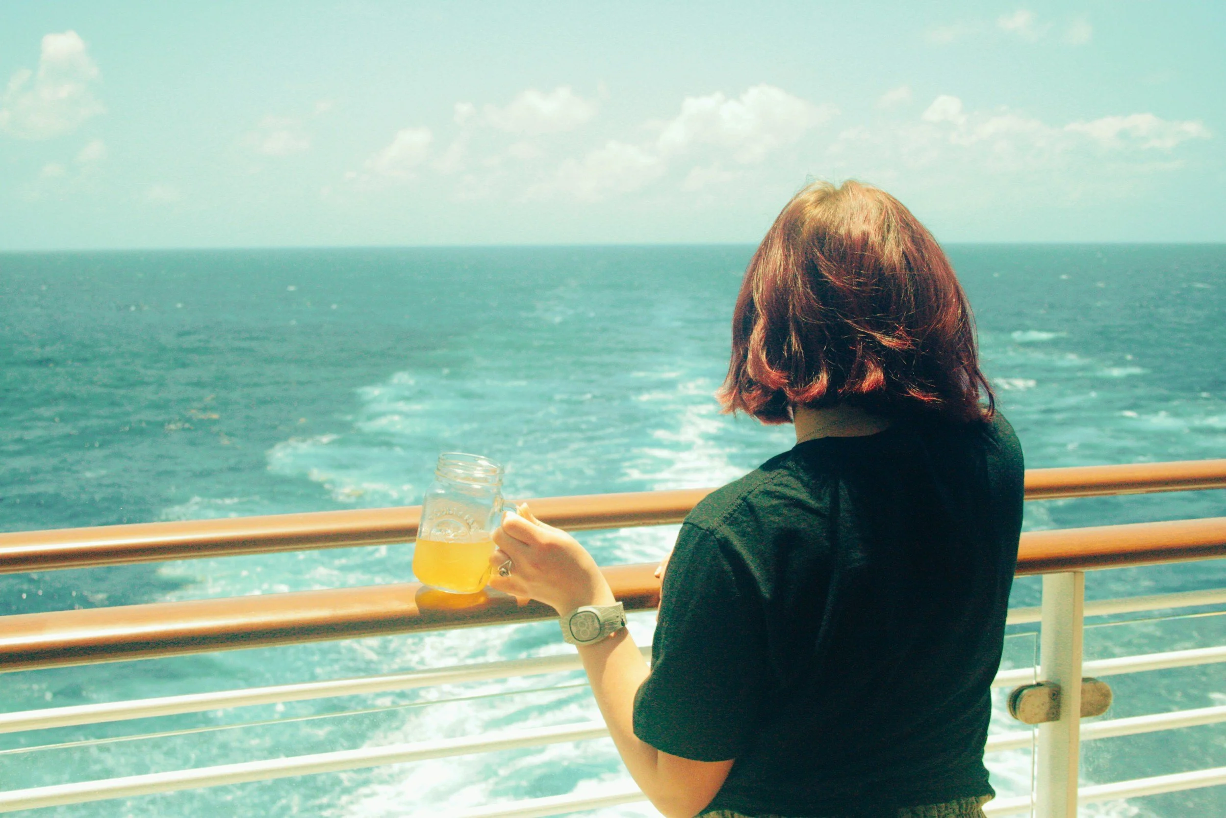 Una donna con capelli corti e rossi si affaccia sulla balaustra di una nave, guardando il mare aperto e bevendo una bibita gialla in un bicchiere di vetro.