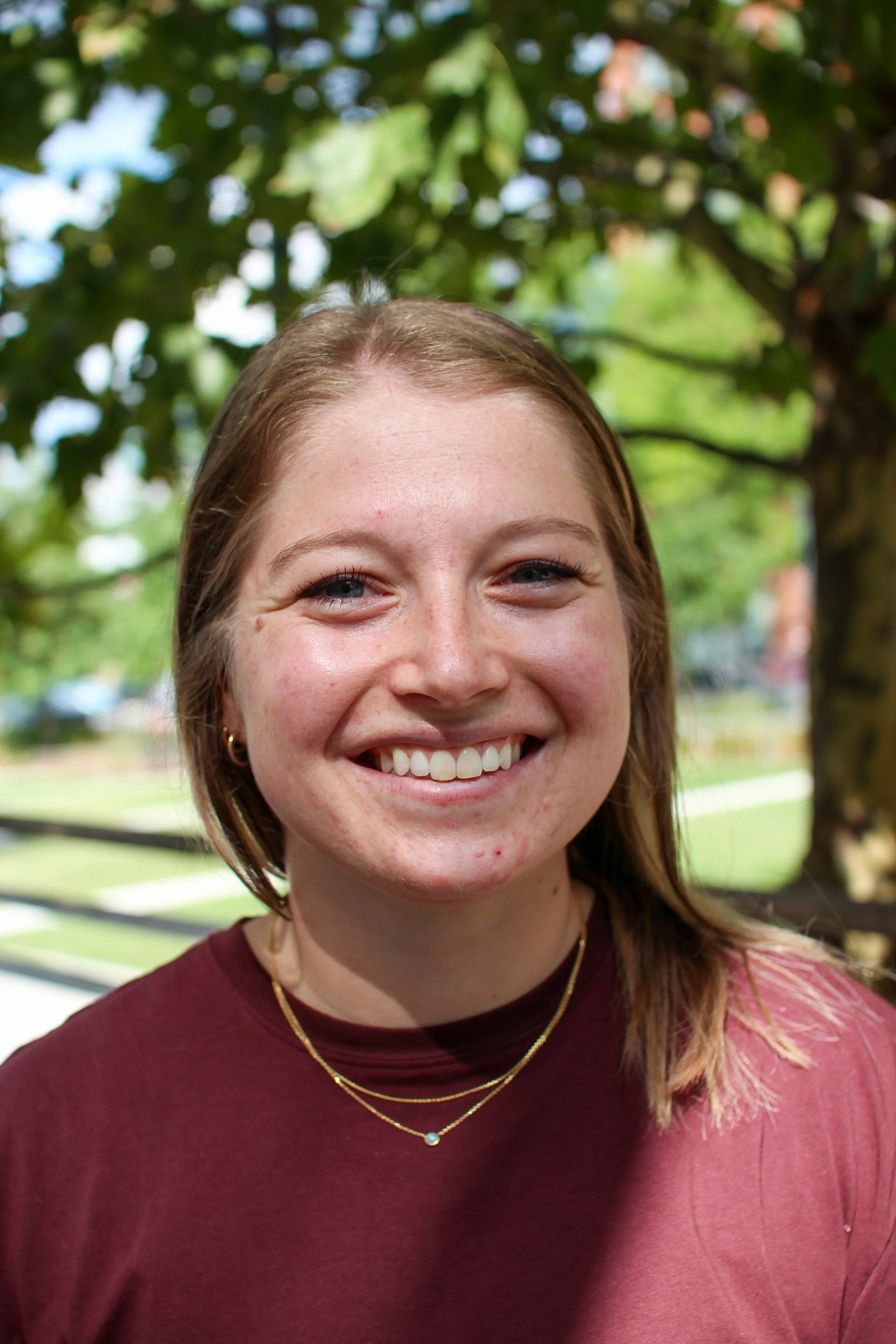 Close-up of a smiling young woman with light brown hair, wearing a maroon shirt and layered gold necklaces, outdoors with green trees in the background.