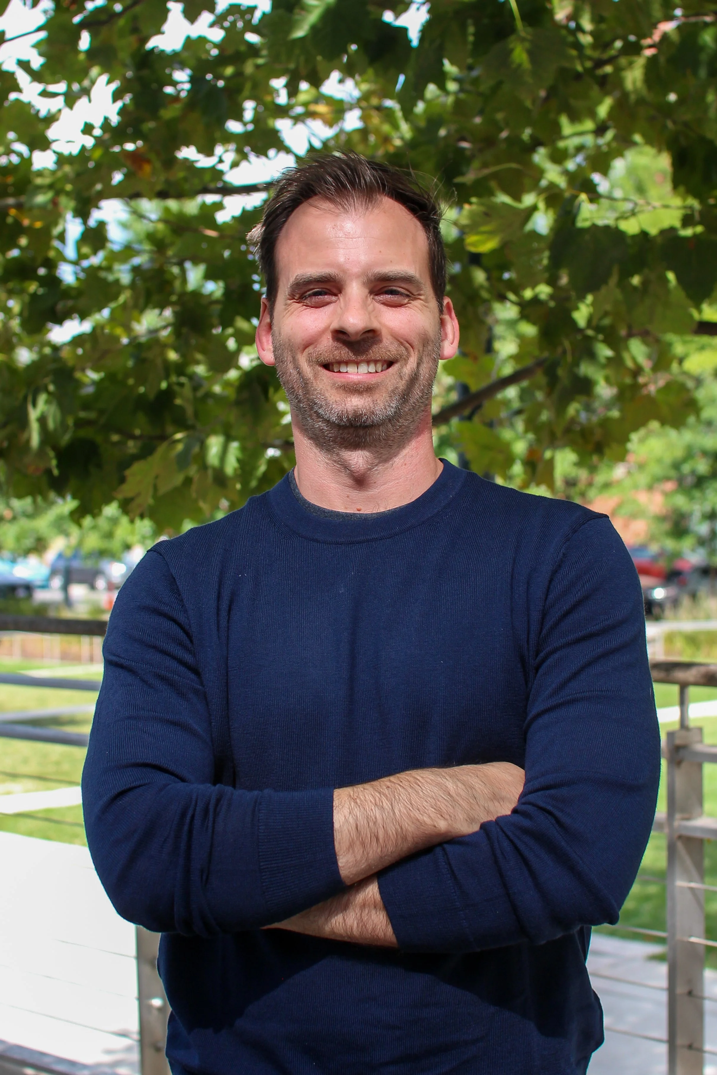 A man with dark hair and a beard, wearing a navy blue long-sleeve shirt, standing outdoors with arms crossed in front of green leafy trees.