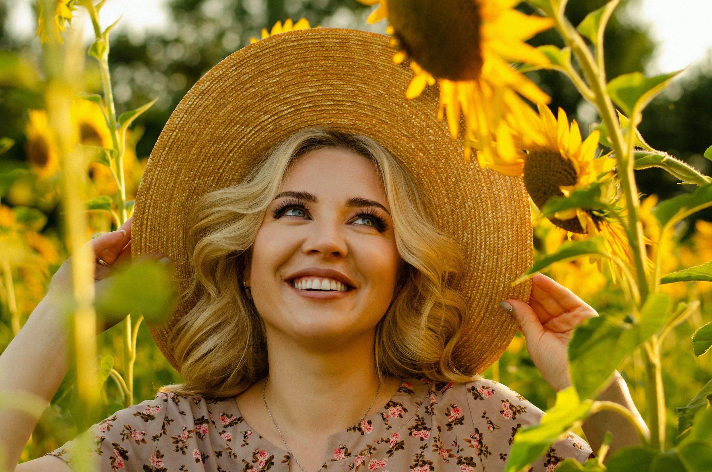 A smiling woman in a floral dress and wide-brimmed straw hat, standing among sunflowers in a field.