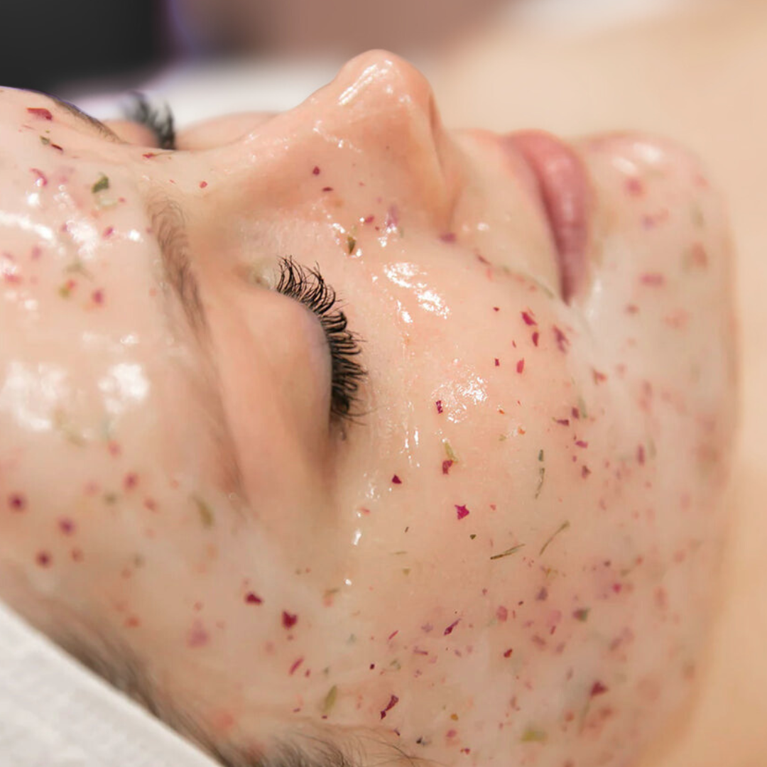 Close-up of a woman's face with skincare mixture containing flower petals, lying down with eyes closed.