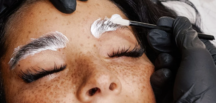 A person with freckles has their eyebrows 'laminated' by a technician applying a clear gel with a brush.