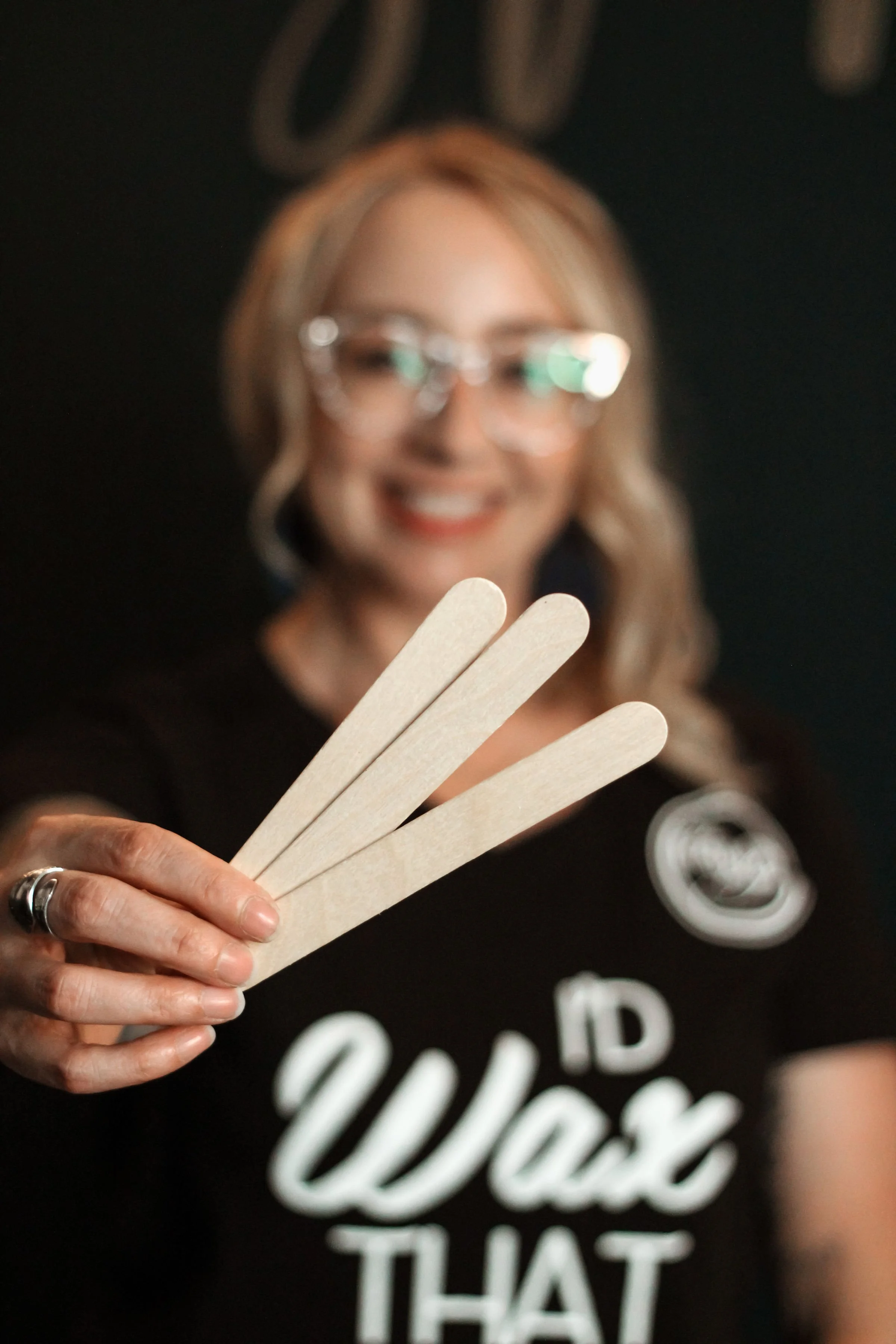 A smiling woman with glasses holds three wooden popsicle sticks toward the camera, with a blurred background.
