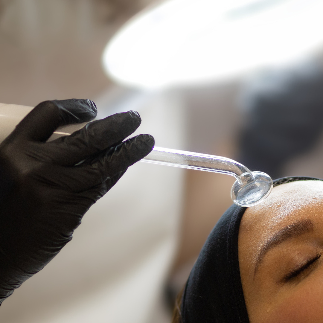 A person receiving a facial treatment with a glass roller on their forehead, wearing a black face mask and black gloves, in a skincare clinic.