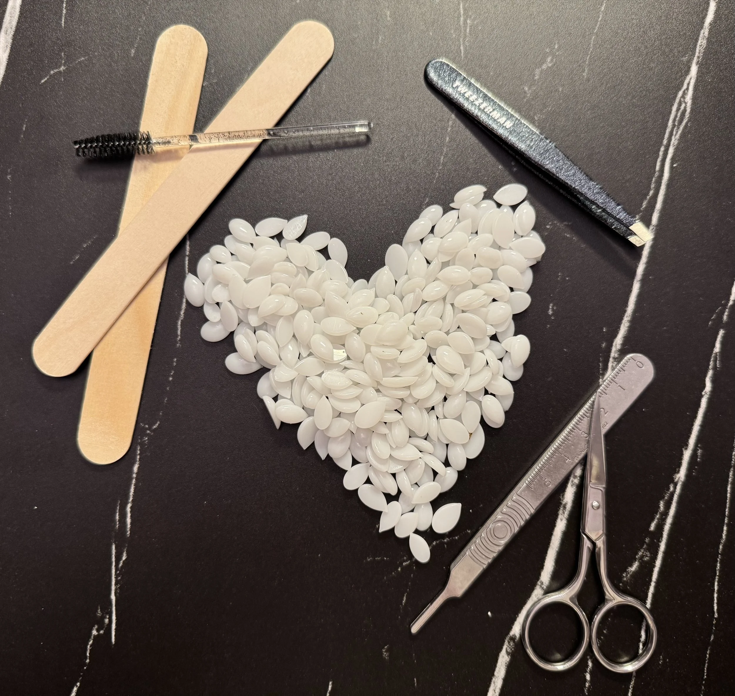 A heart shape made of white nail polish gel beads on a black surface, surrounded by nail care tools including wooden sticks, a metal cuticle pusher, a small scissors, a nail file, and an orange cuticle pusher.