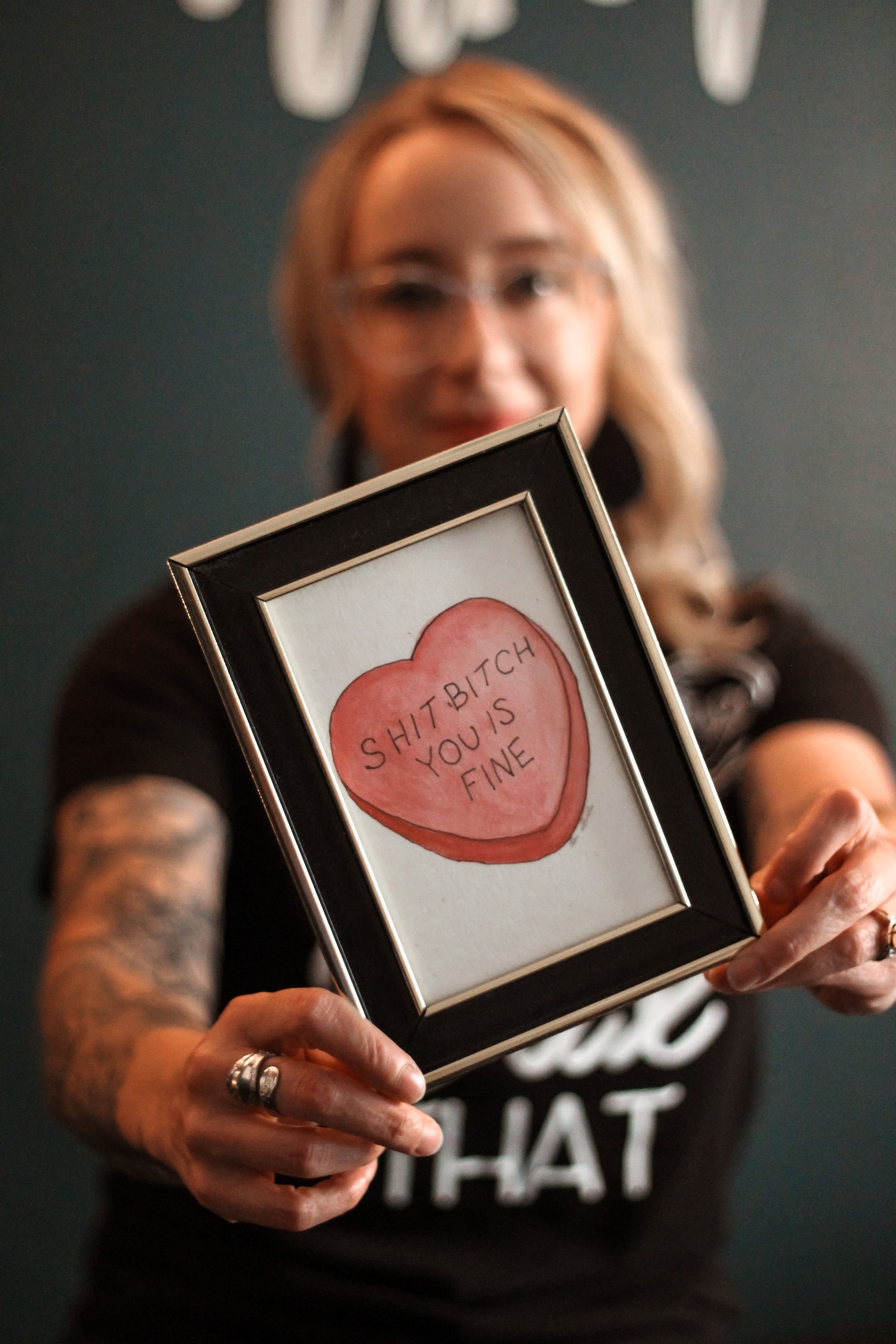 A woman with glasses and tattoos holding a framed drawing of a heart with the words "SHIT BITCH YOU IS FINE" written inside.