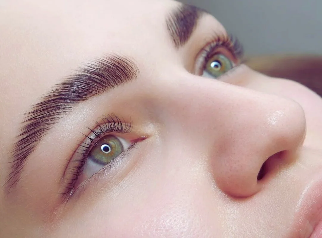 Close-up of a person's face showing well-groomed eyebrows, light makeup, and green eyes with long eyelashes.