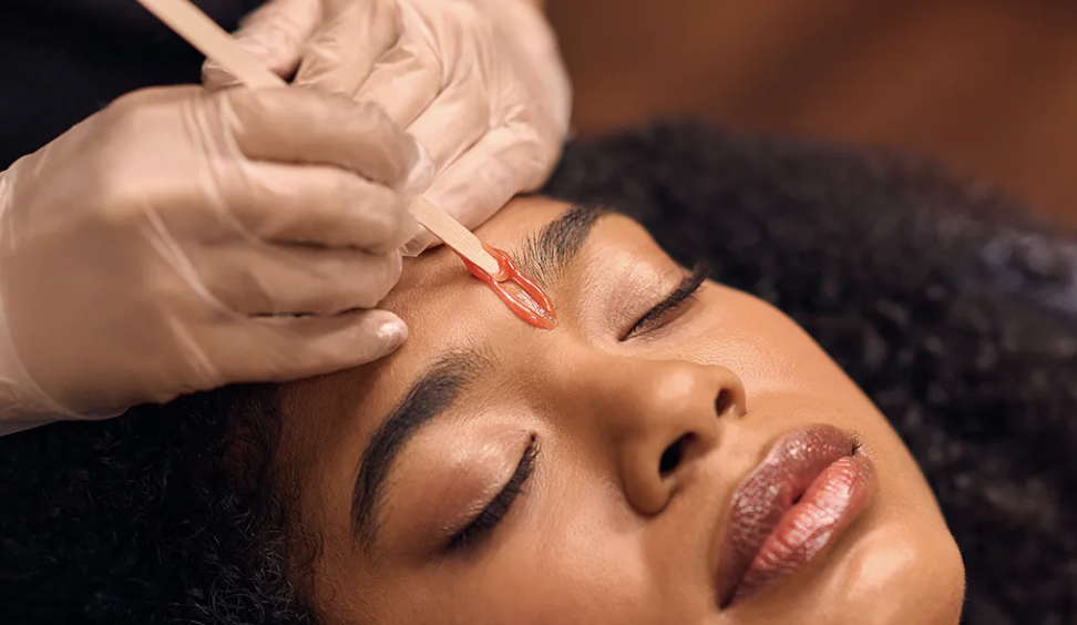 A woman laying down with closed eyes, receiving a eyebrow wax treatment. 