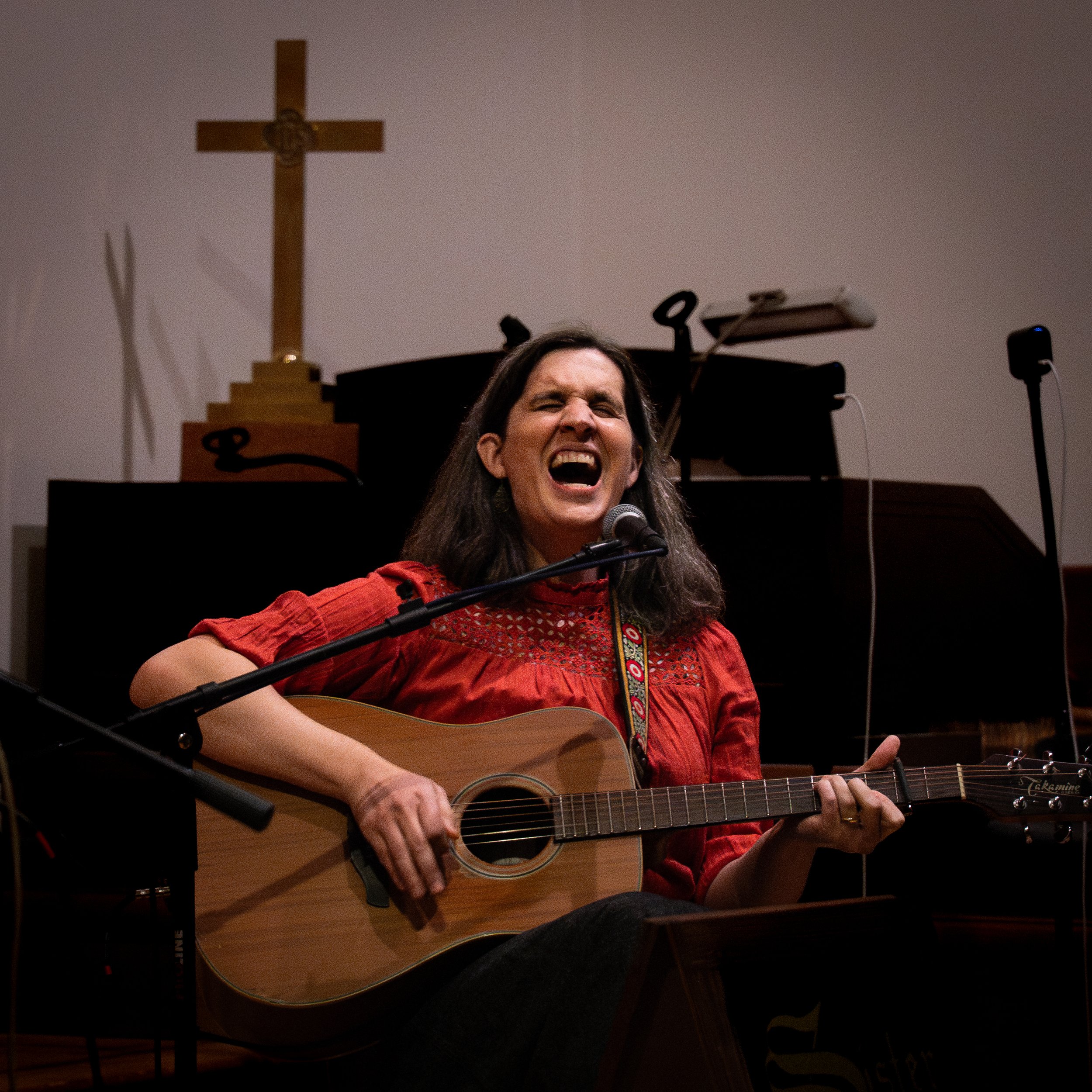 A straight-on shot of Deirdre Jane Rundle singing and playing guitar.