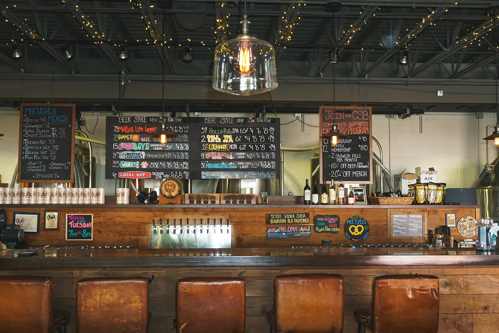 Bar area with chalkboard menus listing beer styles, merchandise, and specials, illuminated by hanging Edison bulbs, with several brown leather chairs in the foreground.