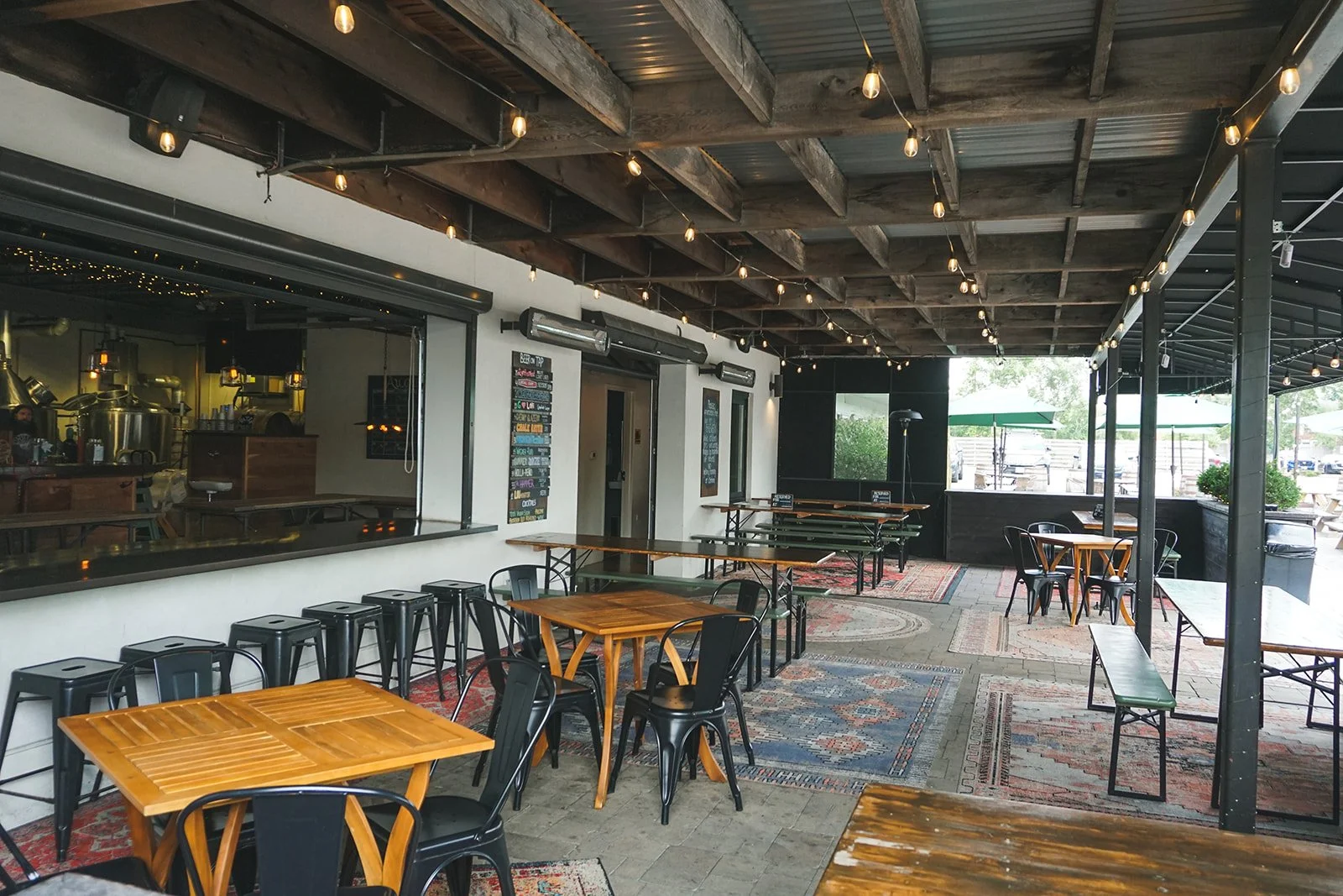 Empty outdoor patio area with wooden tables, black chairs, patterned rugs, and string lights under a wooden ceiling, adjacent to an open kitchen or bar area.
