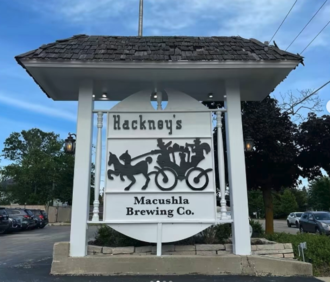 Sign for Hackney's Macushla Brewing Co. with a design of a horse-drawn carriage, on a white background with black text and silhouette, mounted under a building with a shingled roof.