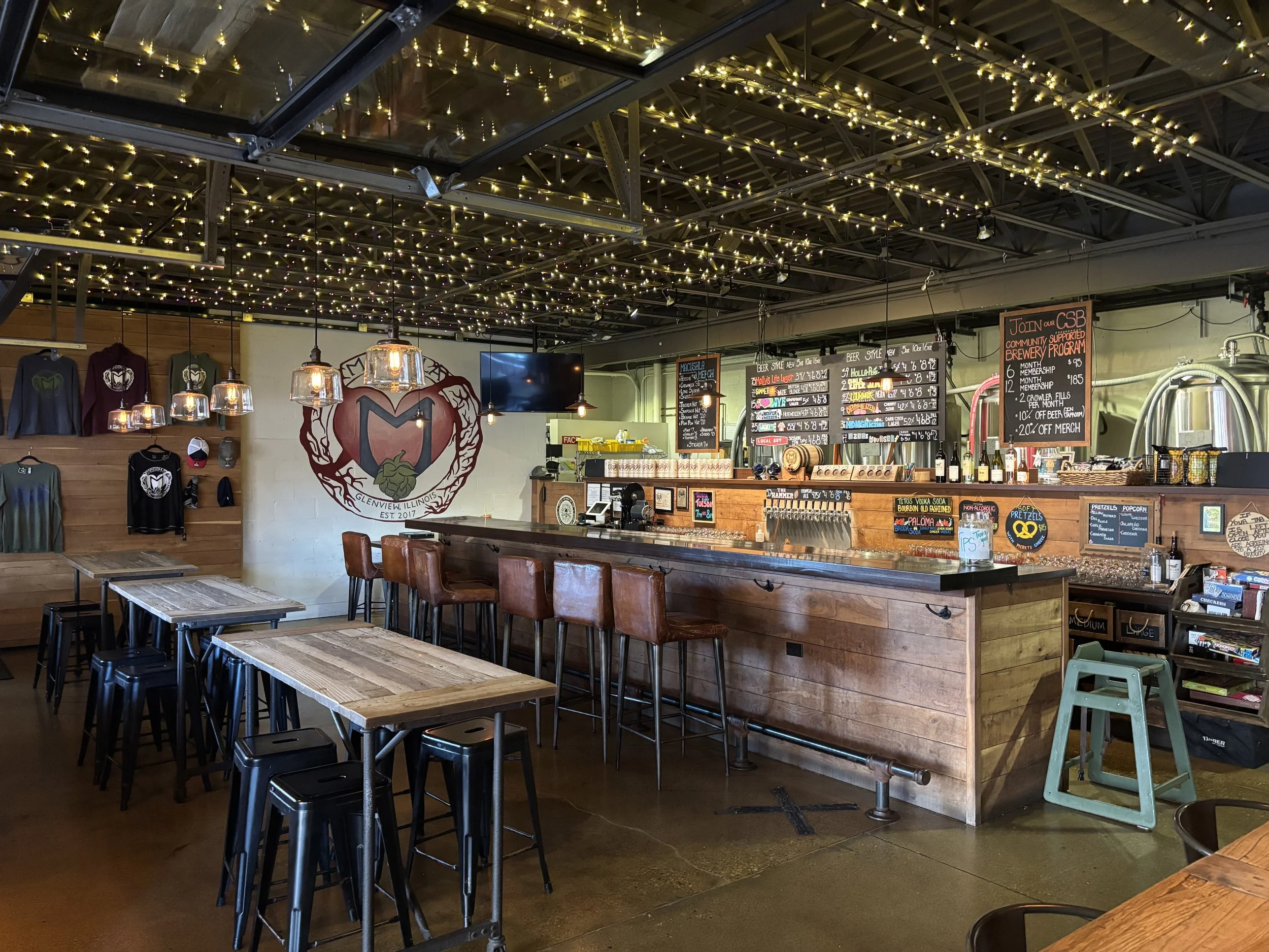 Interior of a brewery bar with string lights hanging from the ceiling, decorated with LED lights. A wooden bar counter with several high chairs. Behind the bar, there's a chalkboard with drink menus and a display with beer bottles. On the wall to the left, there's a logo of a heart with a mountain and the text 'Glenview, Illinois Est. 2017'. Several tables with chairs and stools are in the foreground, alongside wall-mounted clothing and merchandise displays.