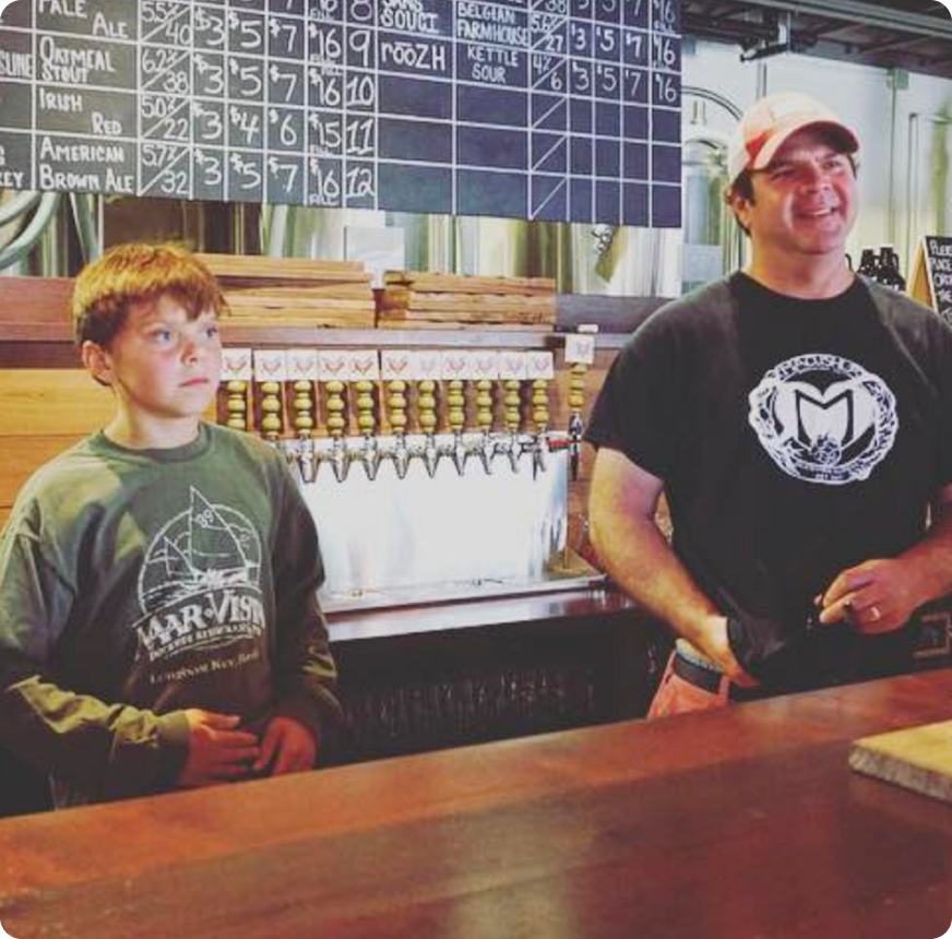 A young boy and an adult man standing behind a bar. The boy is wearing a gray sweatshirt with a boat graphic and the words 'MarVista,' while the man is wearing a black T-shirt with a circular logo and a cap. They are inside a brewery or bar with a chalkboard menu of beers listed above them.
