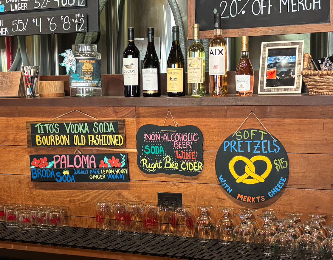 Bar display with bottles of wine on a wooden shelf, colorful handwritten signs advertising Tito's Vodka Soda, Bourbon Old Fashioned, Paloma, Broda's Soda, non-alcoholic drinks, and soft pretzels priced at $15 with details about ingredients and flavors.