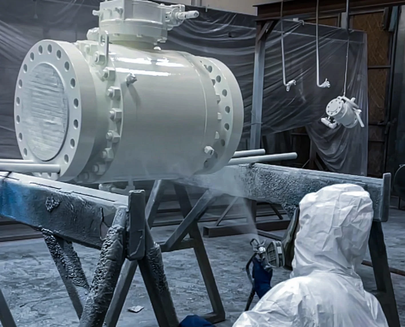 A person in a white protective suit working on industrial painting on a large white cylindrical industrial component in a warehouse or workshop.