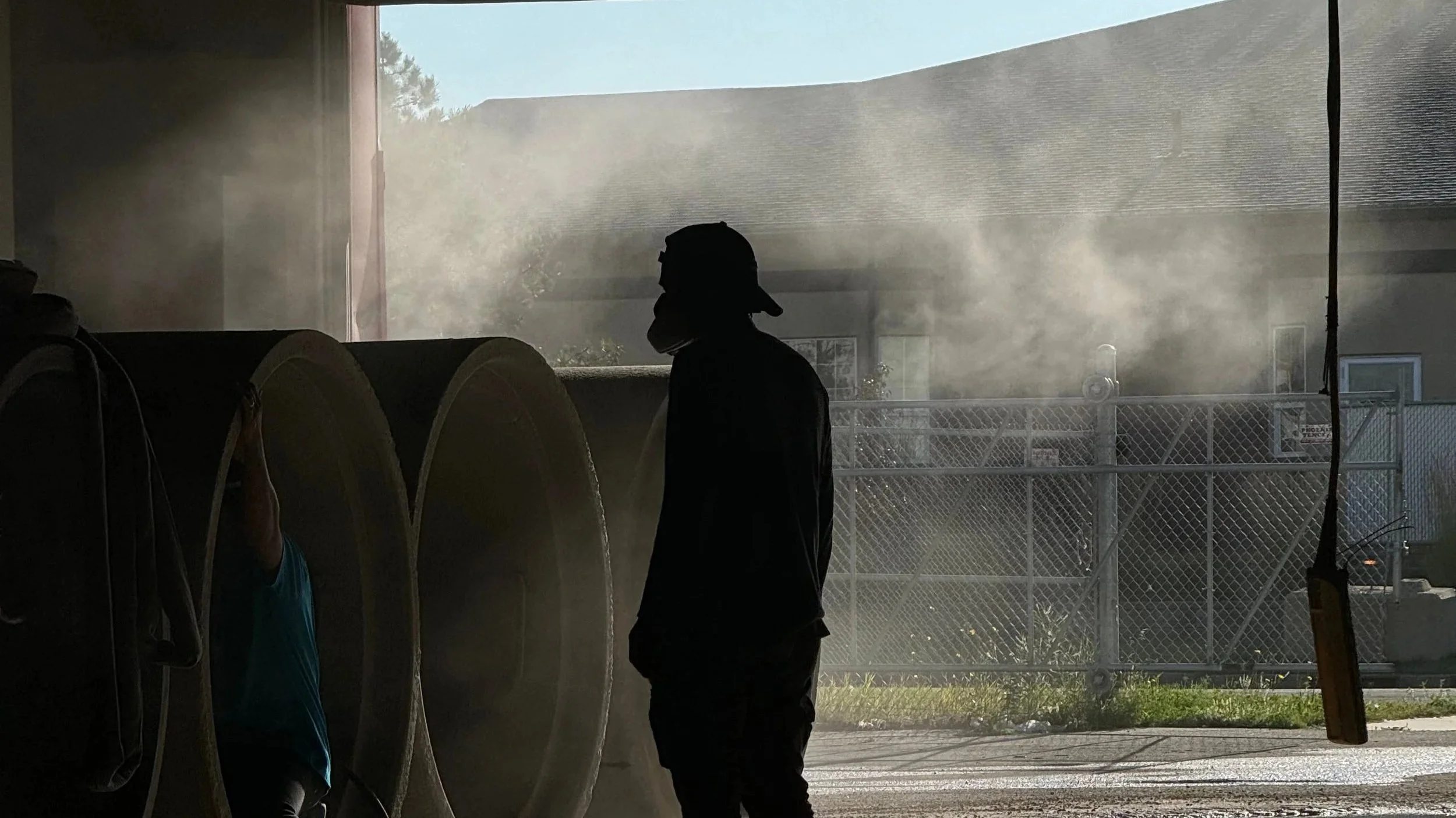 Silhouette of a person wearing a cap and mask standing inside near large pipes where he is Sandblasting, in the background outside is a fenced area.