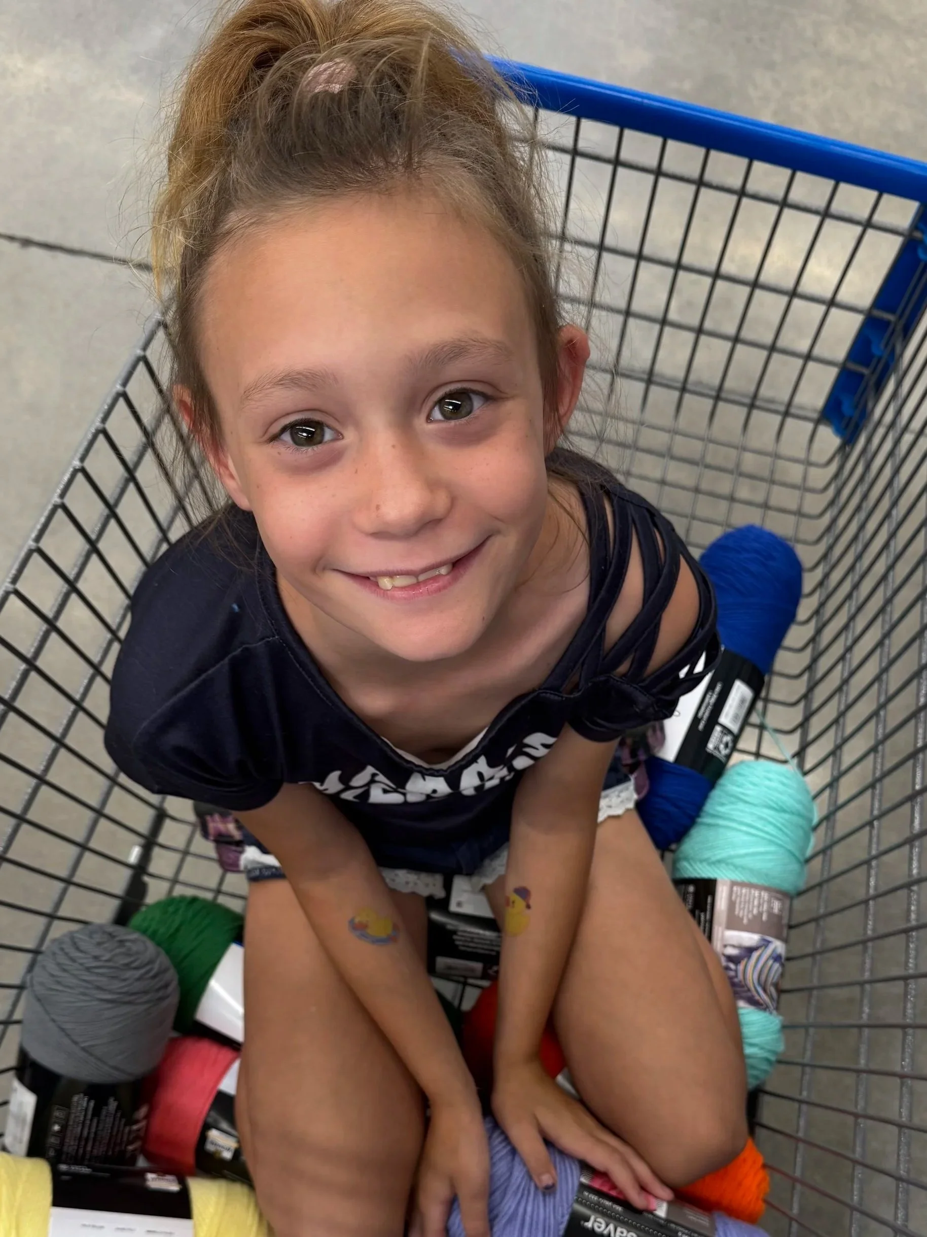A young girl sitting inside a shopping cart filled with colorful yarn. She is smiling up at the camera with a happy expression.