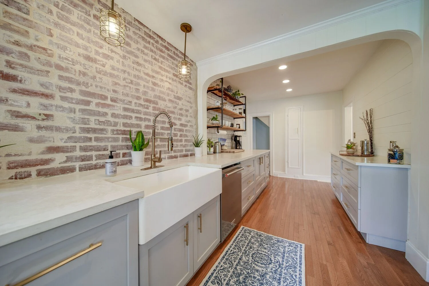 Modern kitchen with white cabinets, a brick accent wall, wooden shelves, plants, and hardwood flooring.
