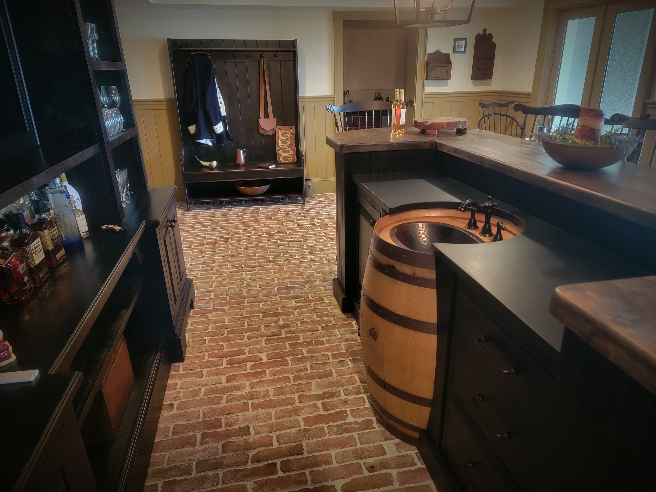 A rustic kitchen with brick flooring, dark wooden cabinets, a small barrel-shaped sink, bottles of alcohol on the side, a wooden stylish coat rack, a window with sunlight, and decorative items on the countertops.