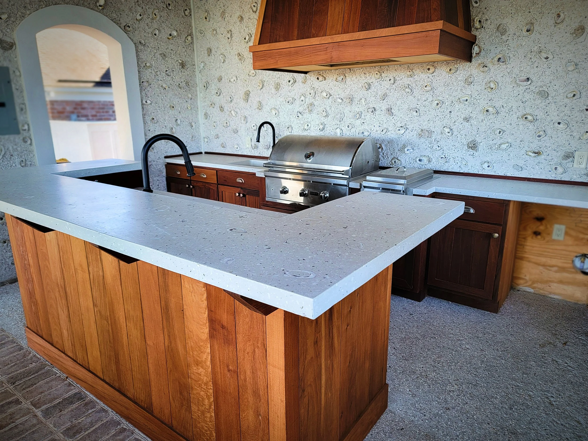 Outdoor kitchen with a white speckled countertop, a built-in stainless steel grill, and black faucets, surrounded by wooden cabinetry and a textured shell stone wall.