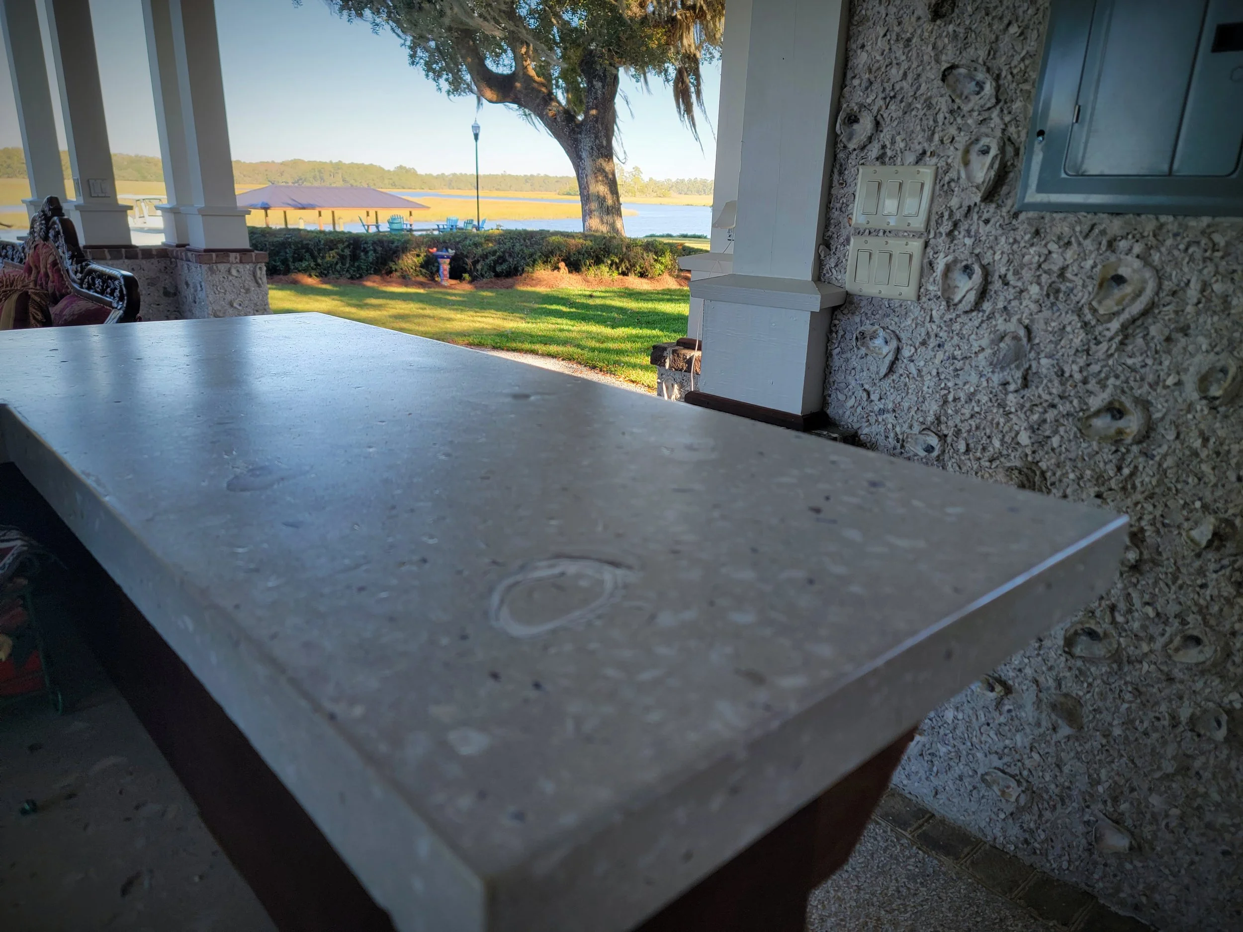 View from a porch or patio showing a concrete countertop, with a grassy area, trees, a tent, and a lake in the background.