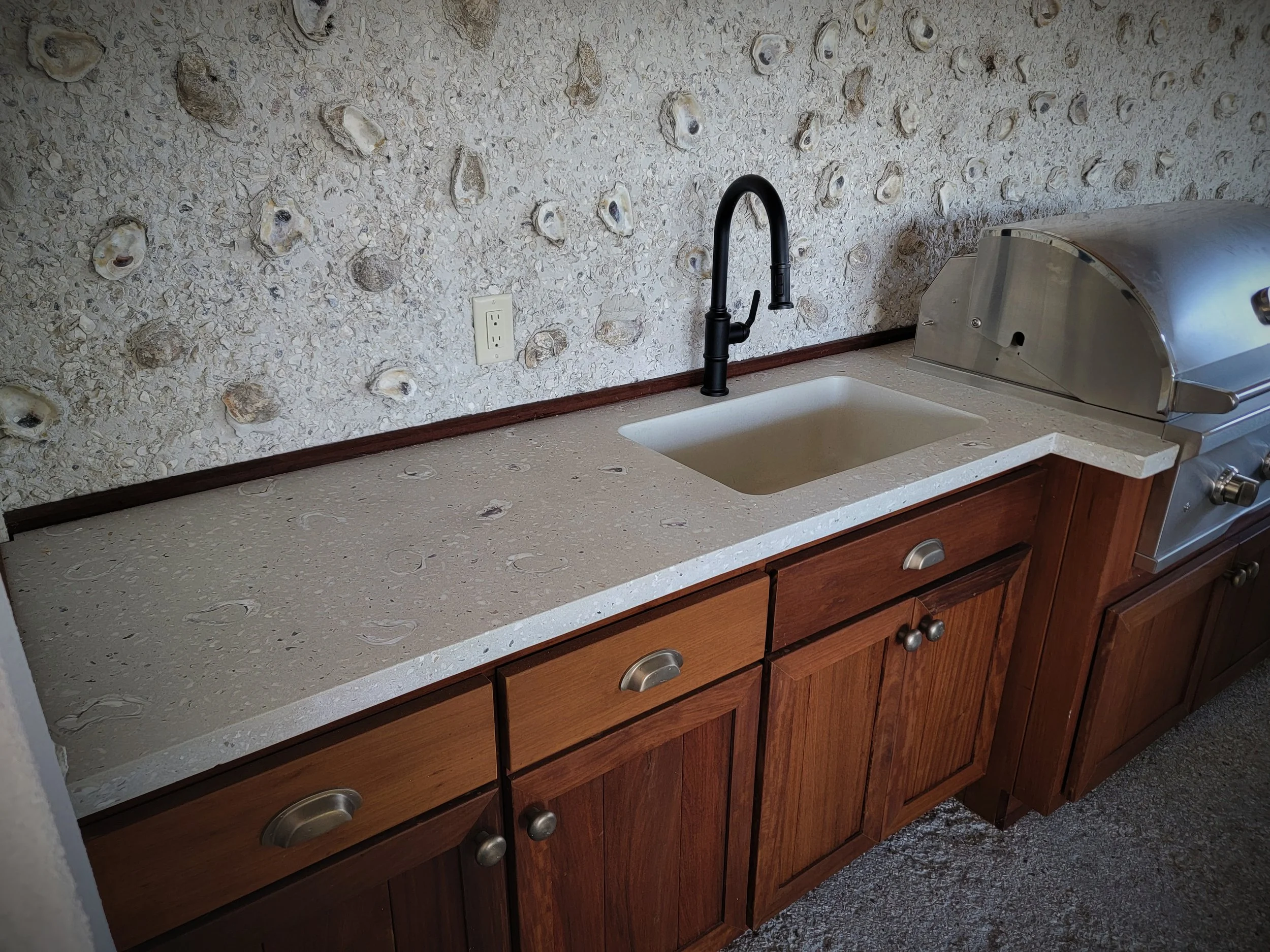 Kitchen countertop with a sink, black faucet, wood cabinets, a wall outlet, and a grill on the right side.