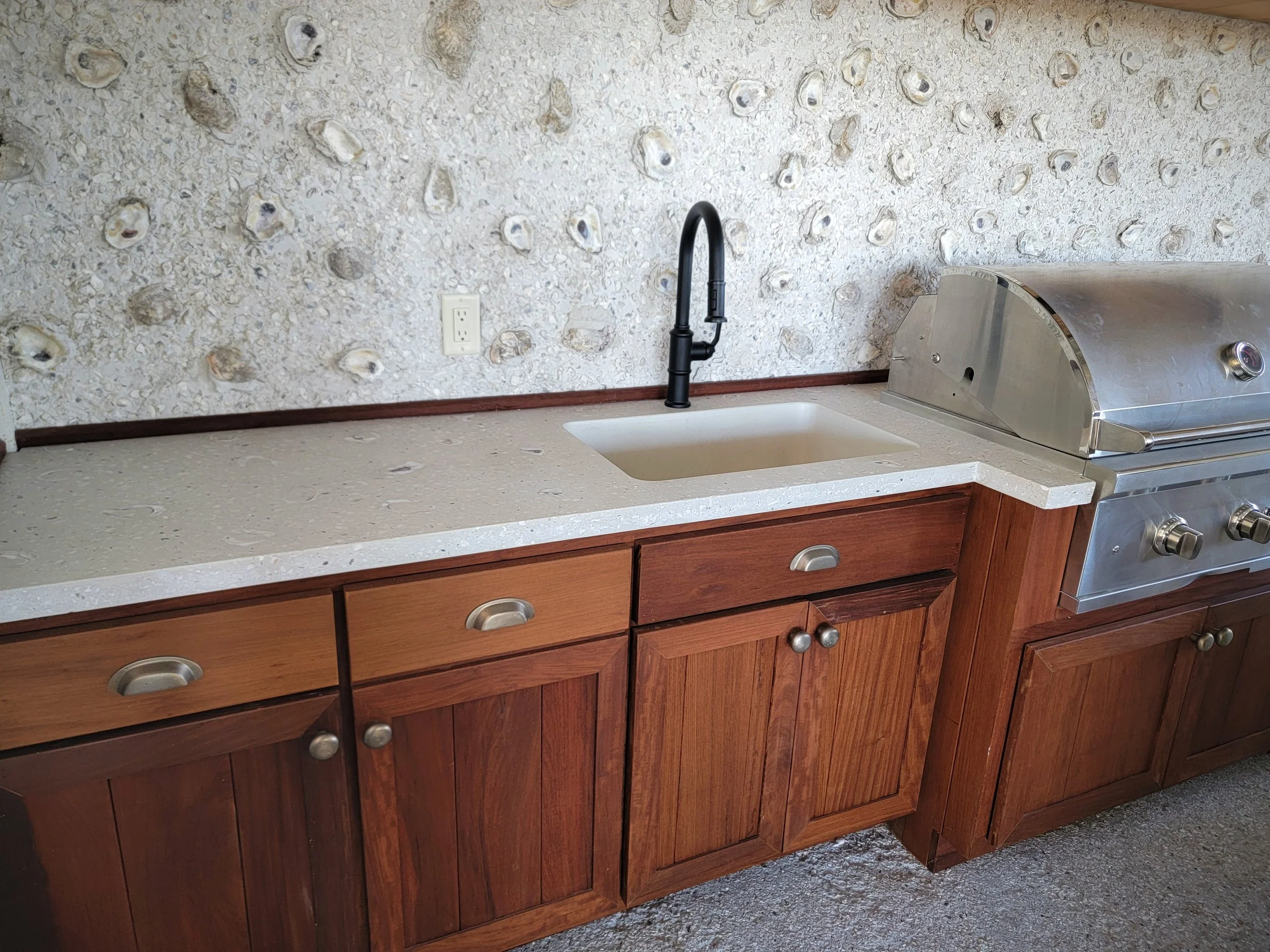Kitchen countertop with a black faucet over a small sink, a stainless steel grill on the right, wooden cabinets below, electrical outlet on the wall, and a textured shell-patterned backsplash.
