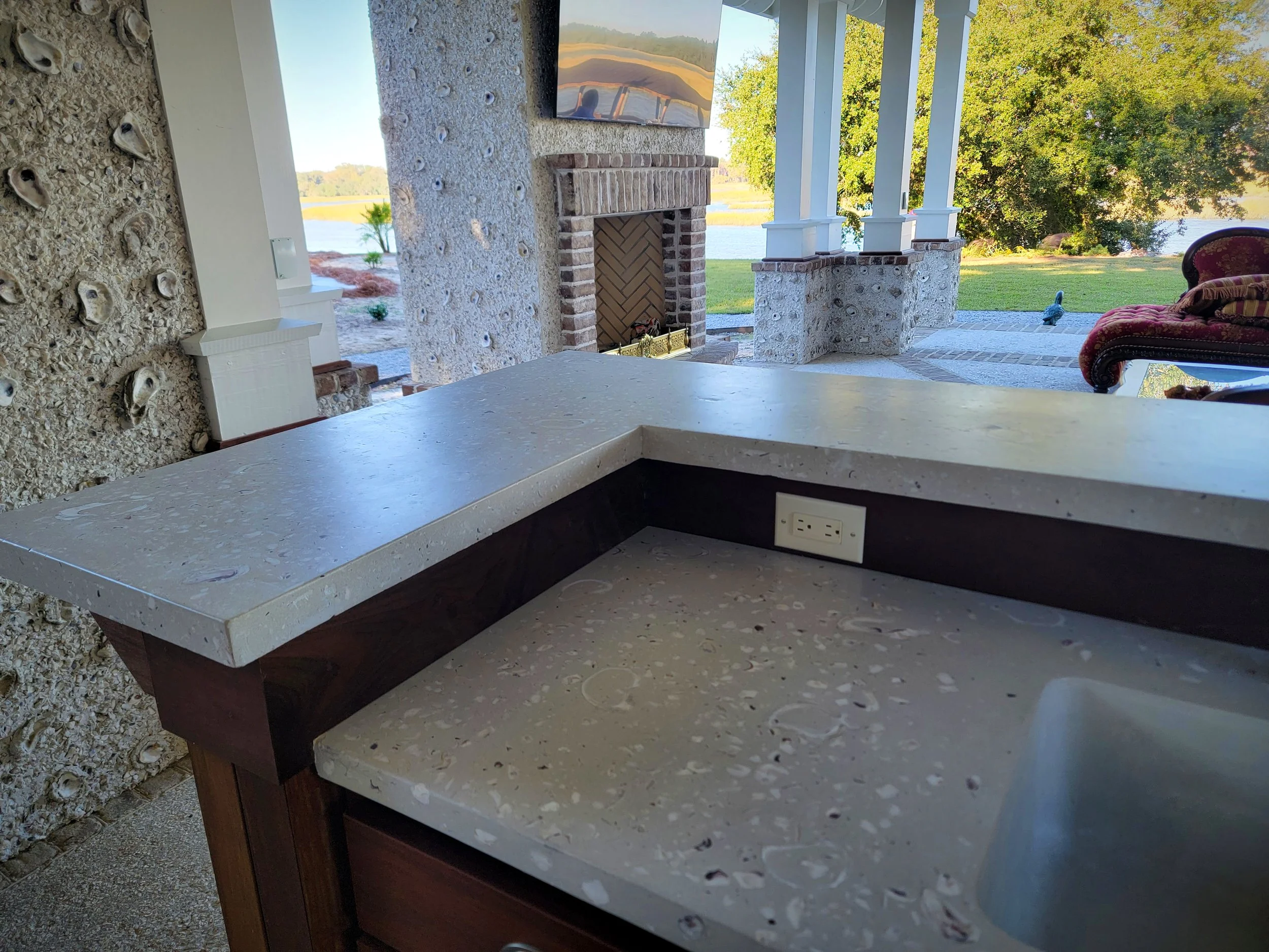 The image shows an outdoor kitchen area with a beige stone countertop, a built-in sink, and an electrical outlet. There is a view of a lake, trees, and a grassy yard with a brick fireplace and columns in the background.