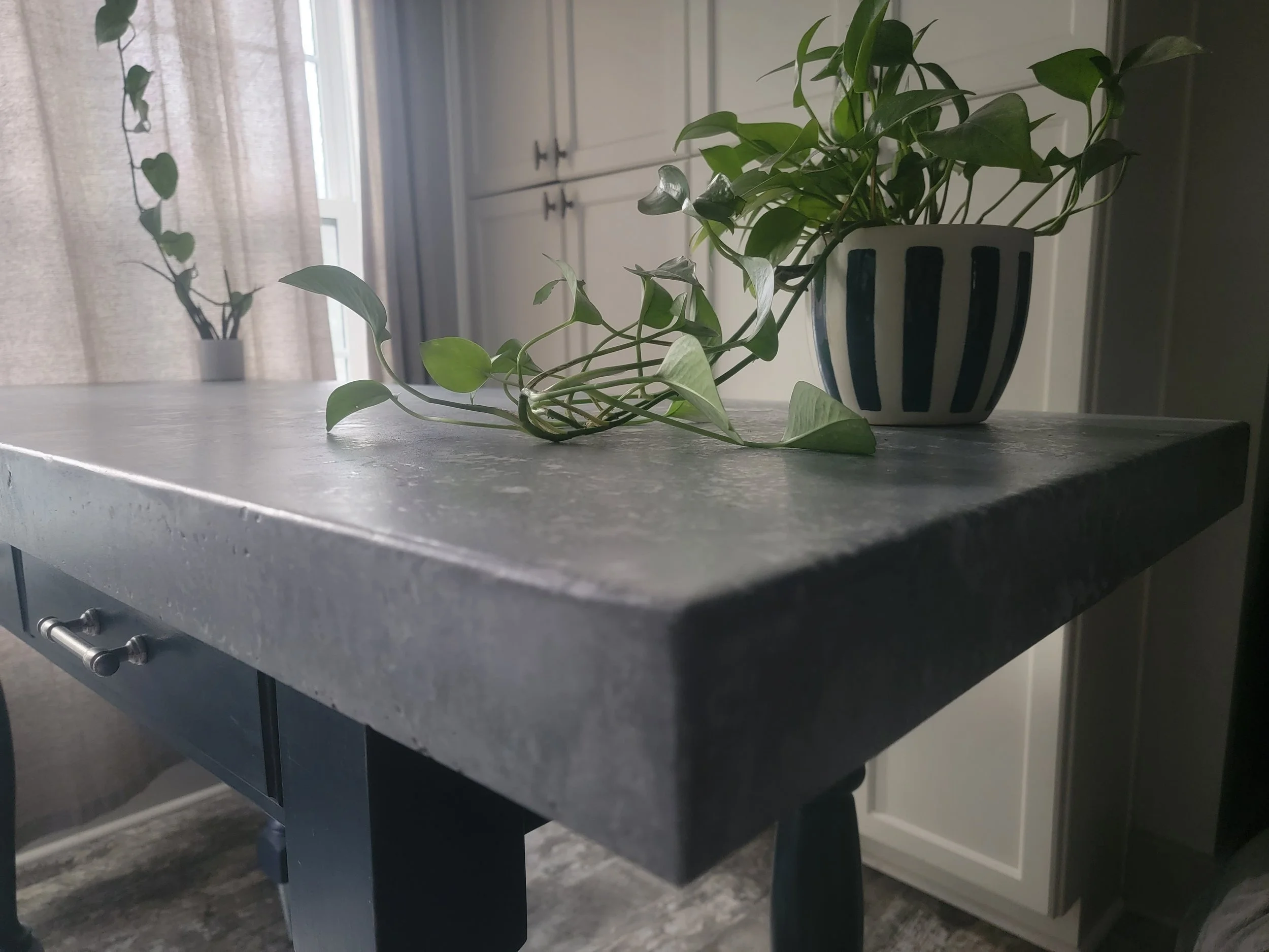 A kitchen countertop with a striped blue and white ceramic pot containing a green leafy plant, and a trailing vine plant in a gray pot in the background near a window with curtains.