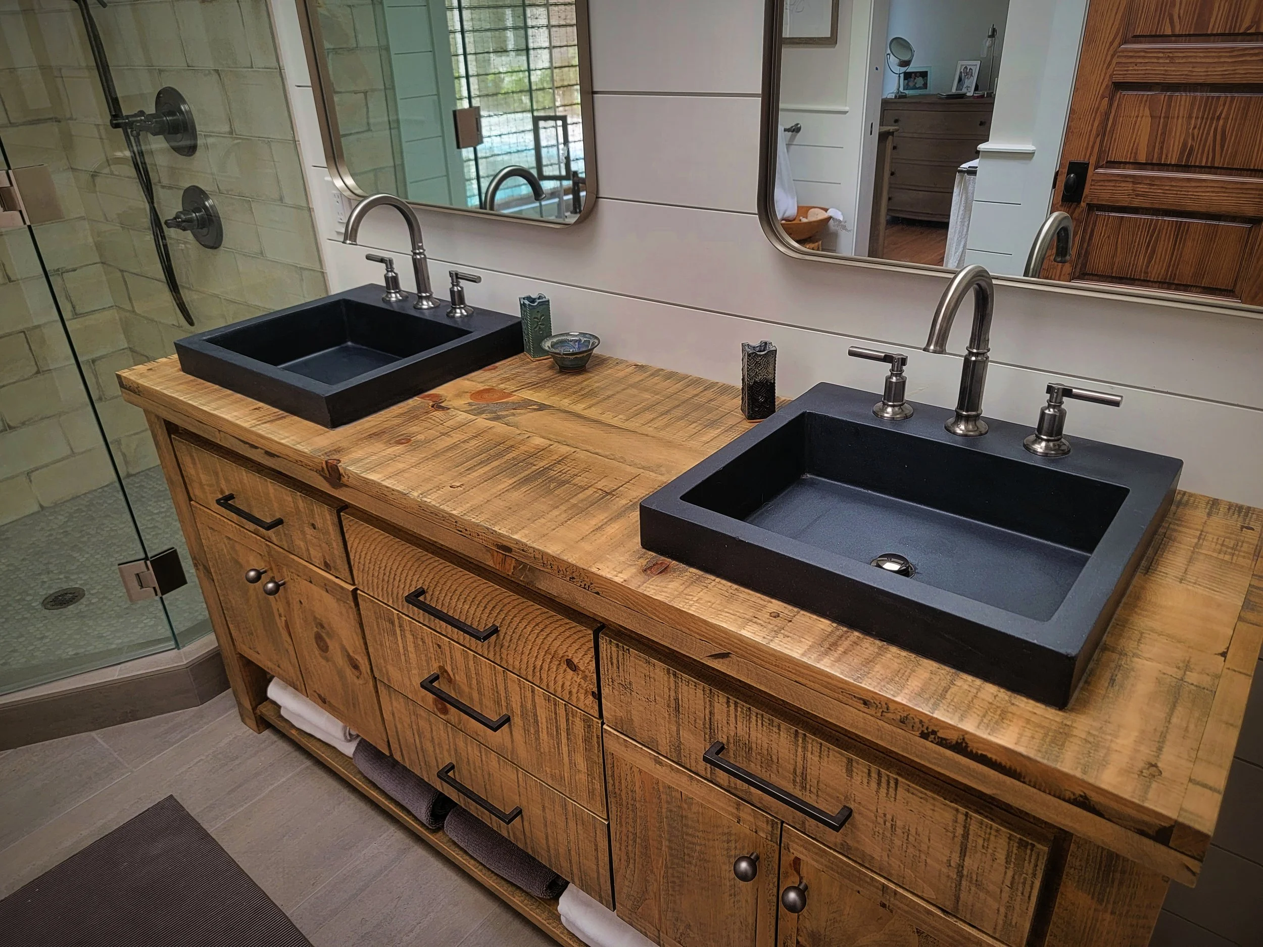 Dual black sinks with silver faucets on a rustic wooden bathroom vanity with mirrors, a shower enclosure, and a view into a bedroom.