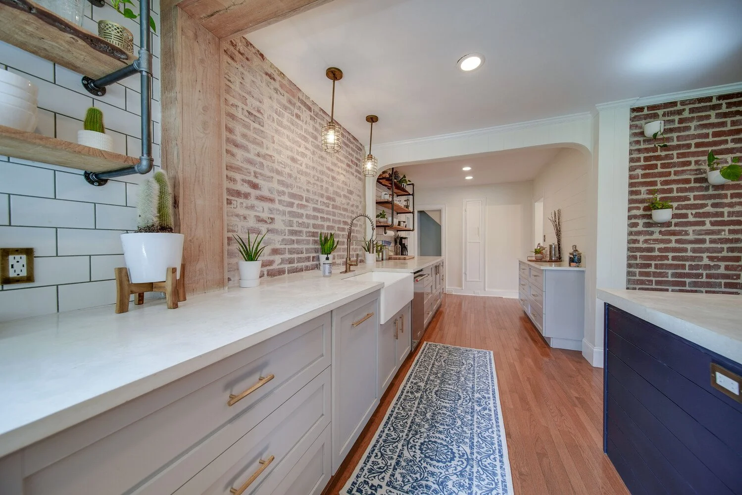 Modern kitchen with white cabinets, brick wall accents, wooden open shelves, a white farmhouse sink, and a decorative blue and white rug on a hardwood floor. Potted plants are placed on the countertops and shelves, with pendant lights hanging from th