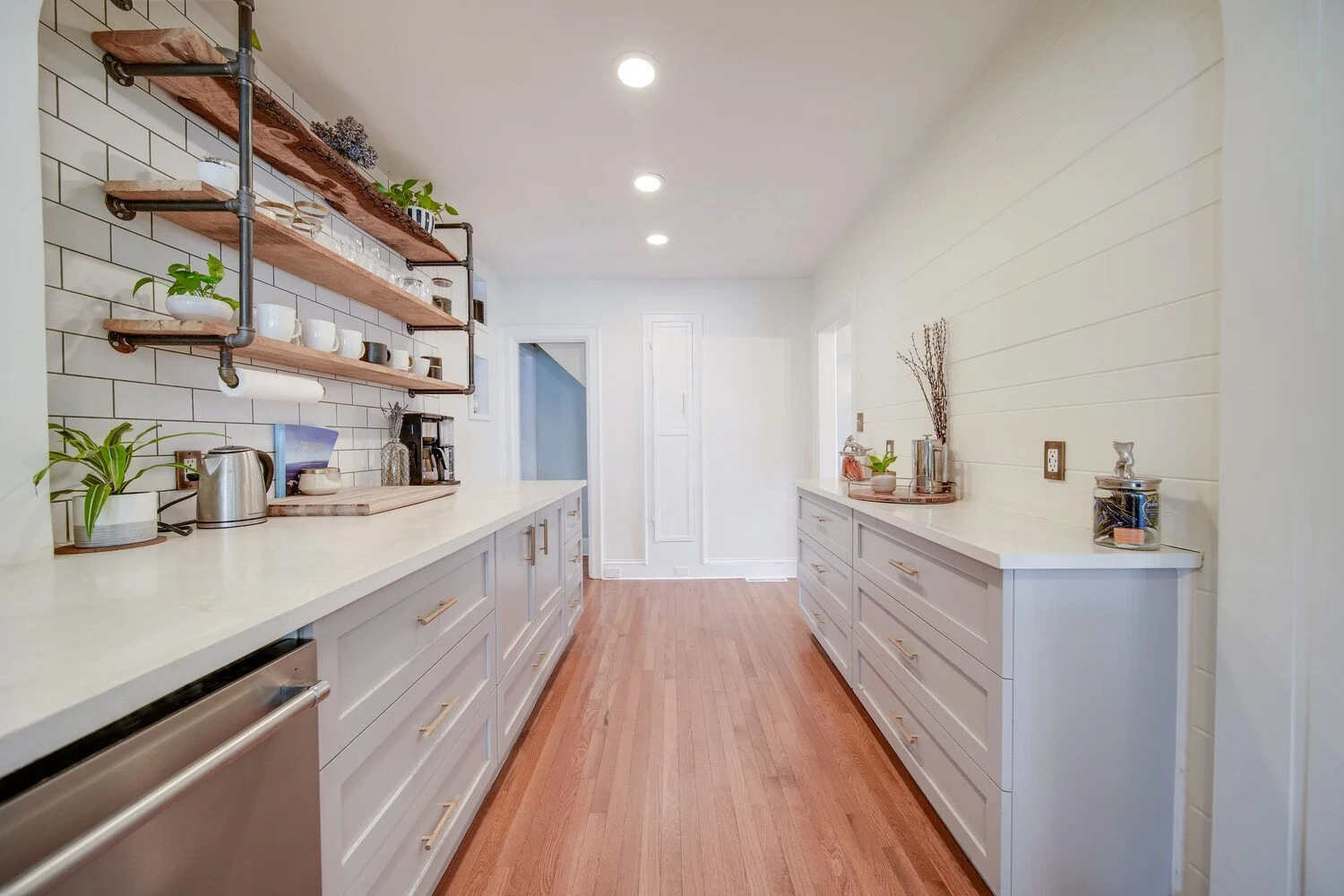 Kitchen with white cabinets, wooden shelves, subway tile backsplash, and hardwood floors. Decor includes plants, cups, jars, and kitchen appliances.