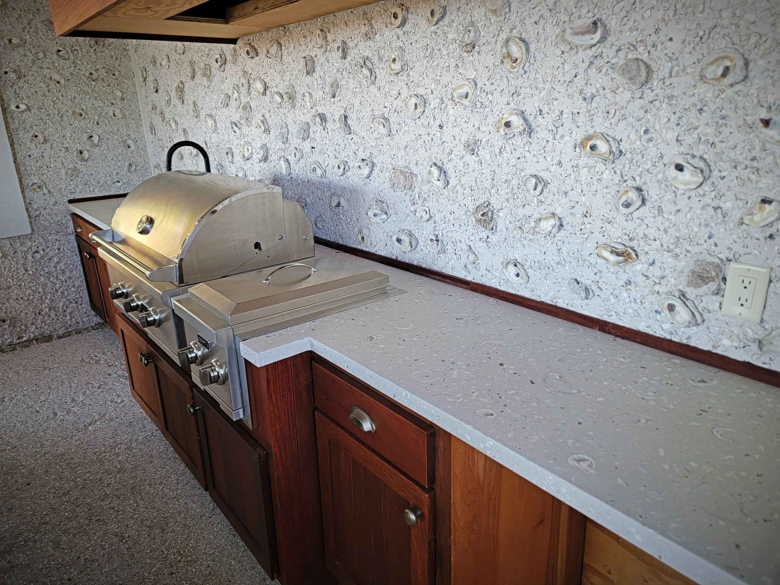 Stainless steel barbecue grill on a kitchen countertop with a wood cabinet below, against a wall with seashell-patterned wallpaper.