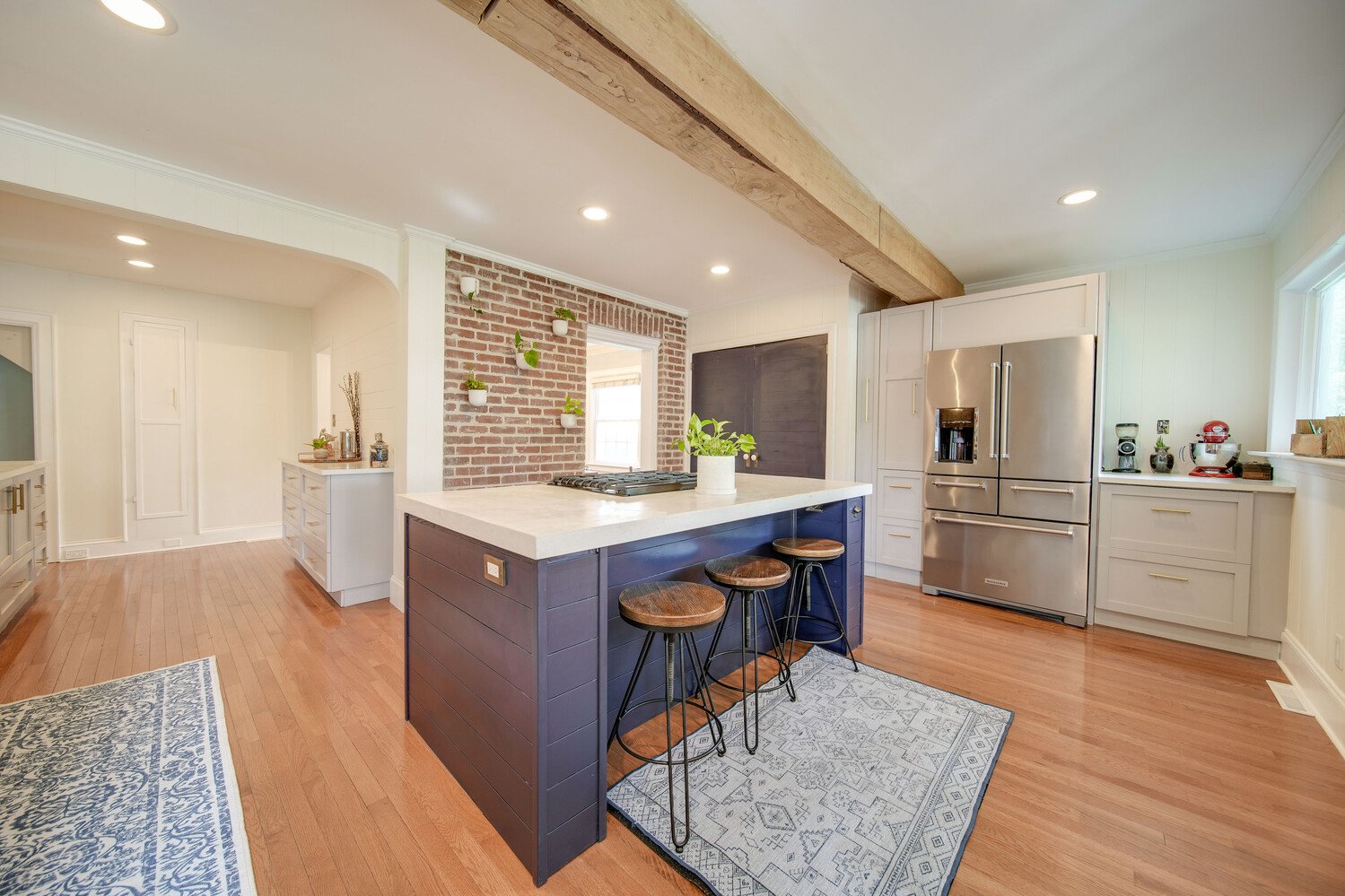 Modern kitchen with stainless steel refrigerator, white cabinets, and a navy blue kitchen island with a white marble countertop. Wooden barstools are at the island, and a small rug is in front of it. There's a brick accent wall with small potted plan