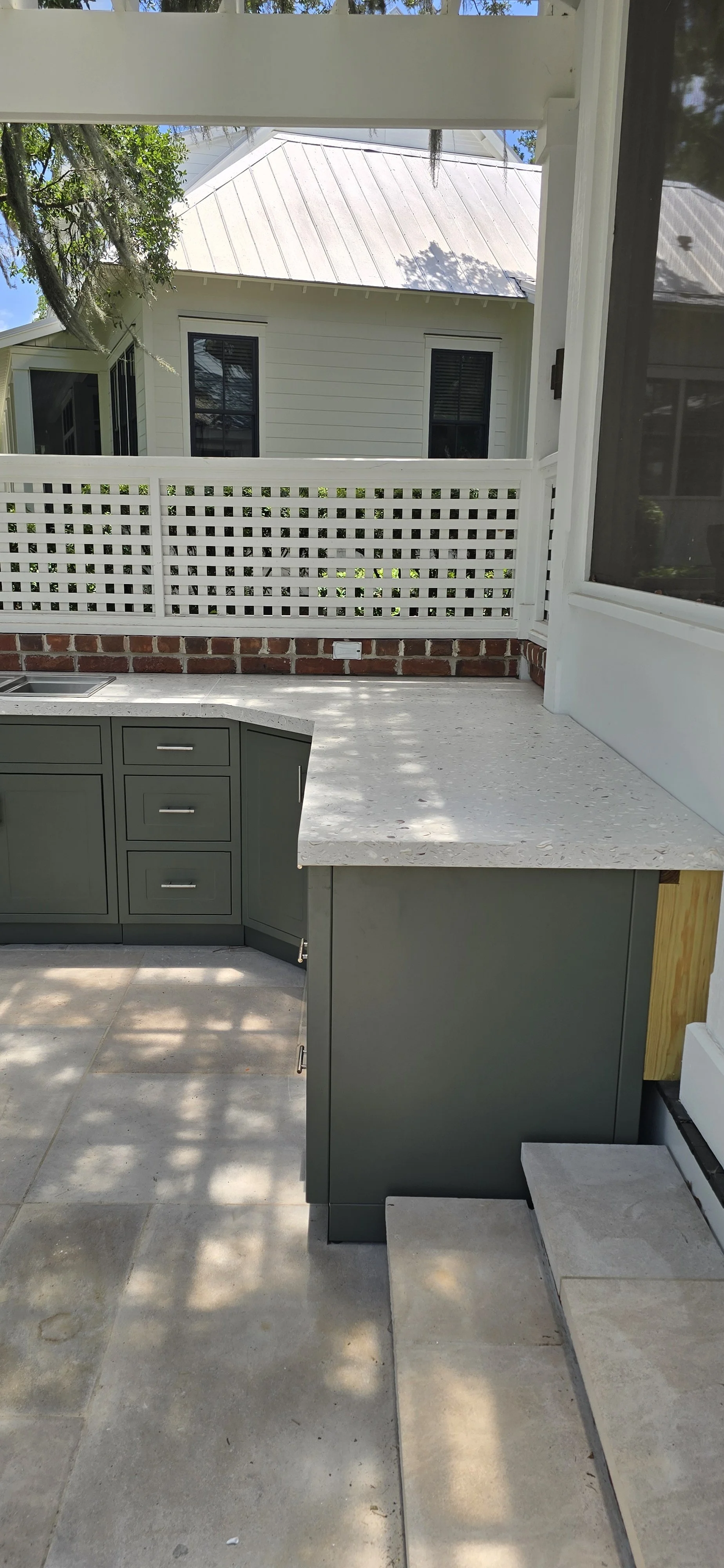 Light concrete outdoor kitchen countertop and deep return above gray cabinetry beneath a covered porch