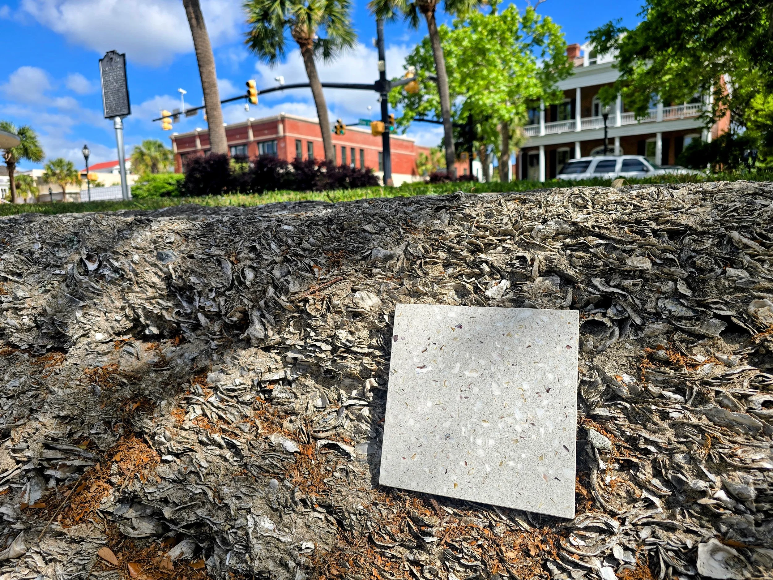 Close-up of a rough stone surface with a small white tile leaning against it. In the background, there are trees, a building, some cars, and traffic lights under a partly cloudy sky.