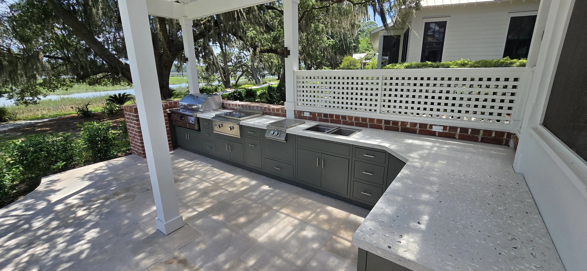 Covered outdoor kitchen with light concrete countertops, grill, sink and a long return over gray cabinetry