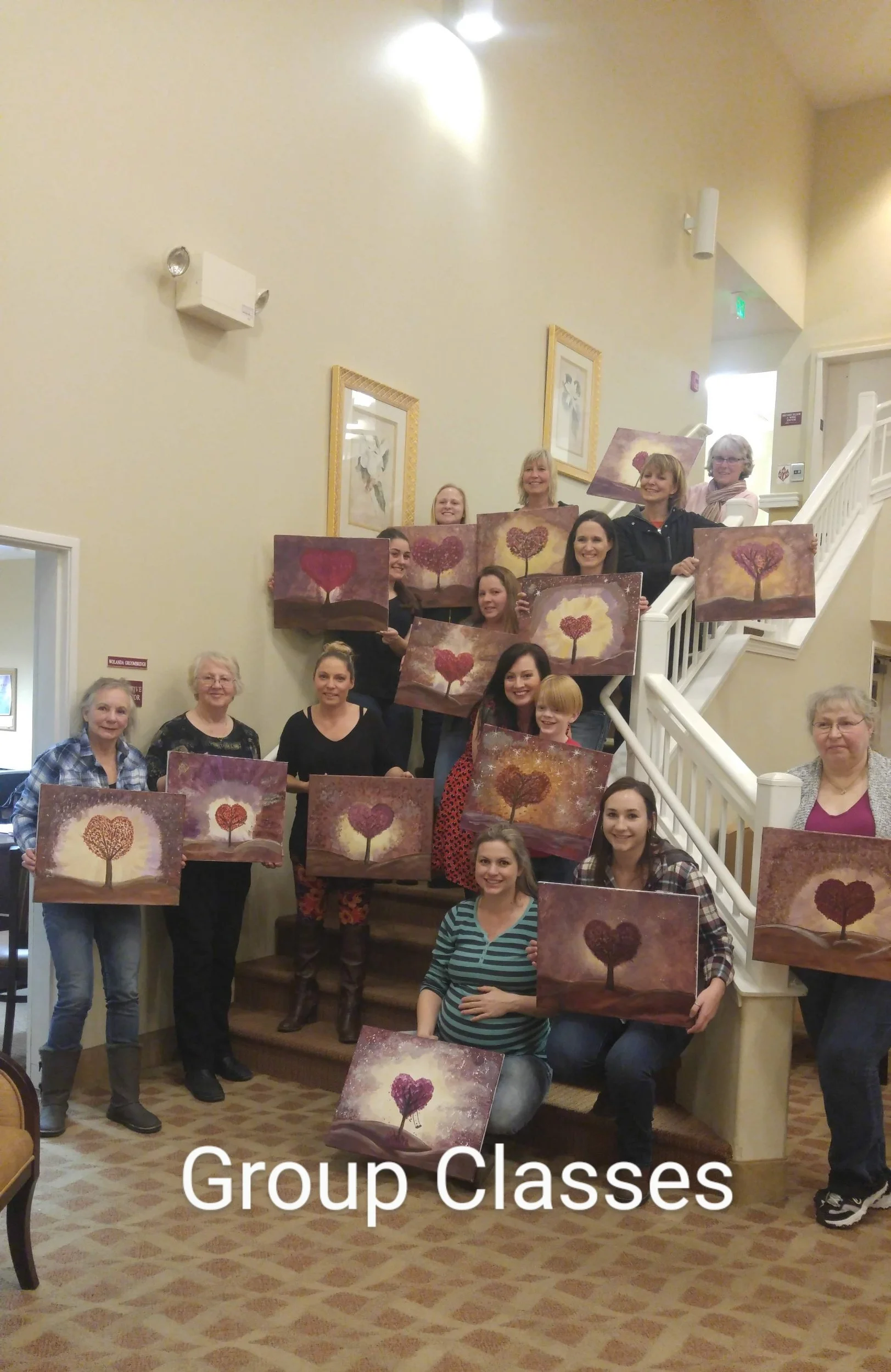 Group of women and a girl posing on staircase, holding paintings of stylized trees with heart-shaped foliage, after a painting class led by June Lizotte Art.