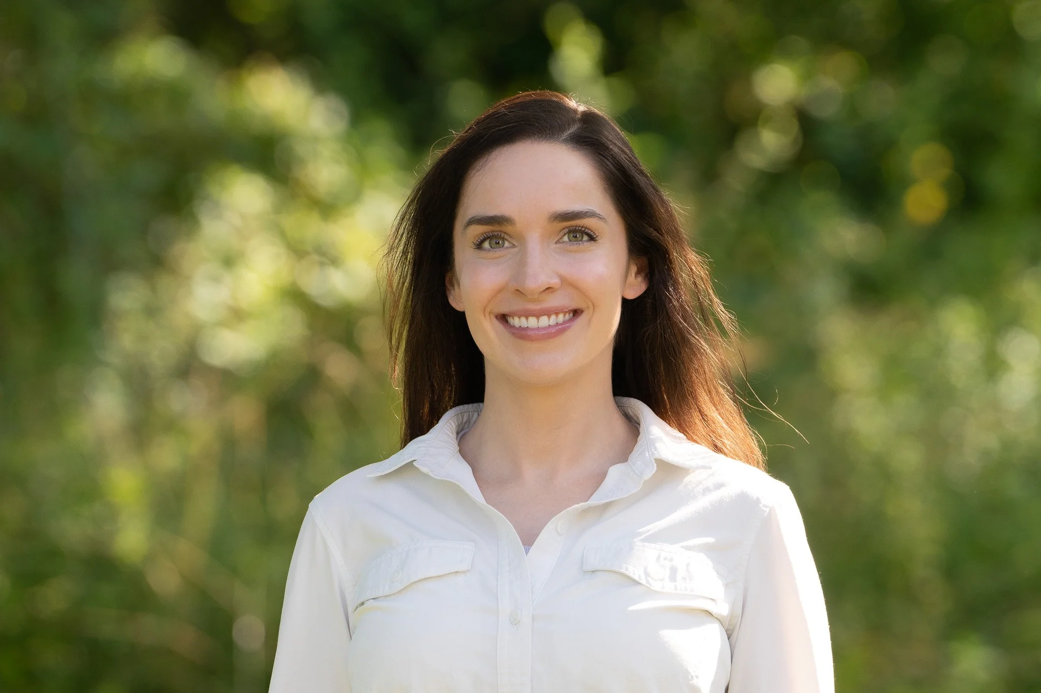 A young woman with long brown hair, fair skin, and green eyes smiling outdoors in front of a blurred green background.