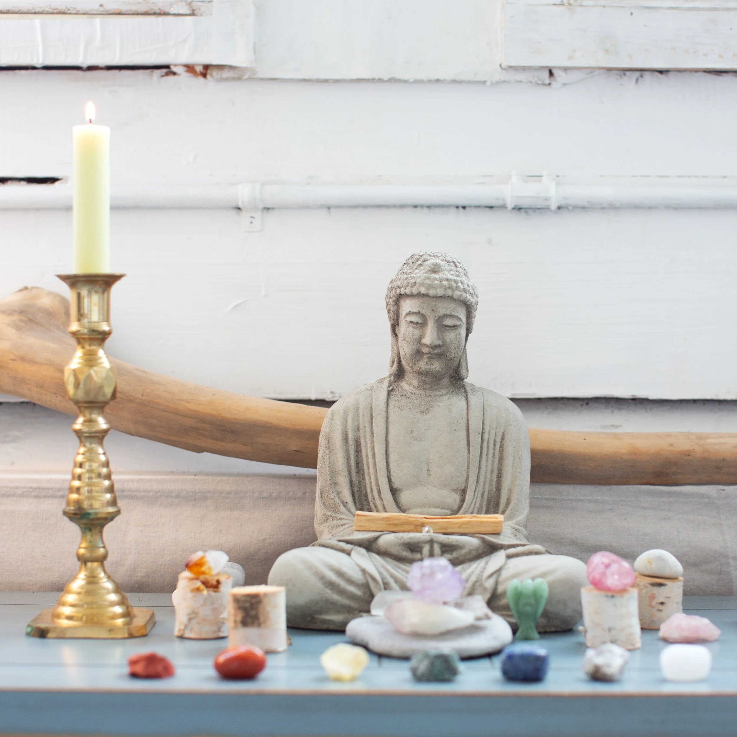 A Buddha statue sitting cross-legged on a ledge surrounded by colorful crystals, with a tall candle on a brass holder to the left and a natural wood branch behind.