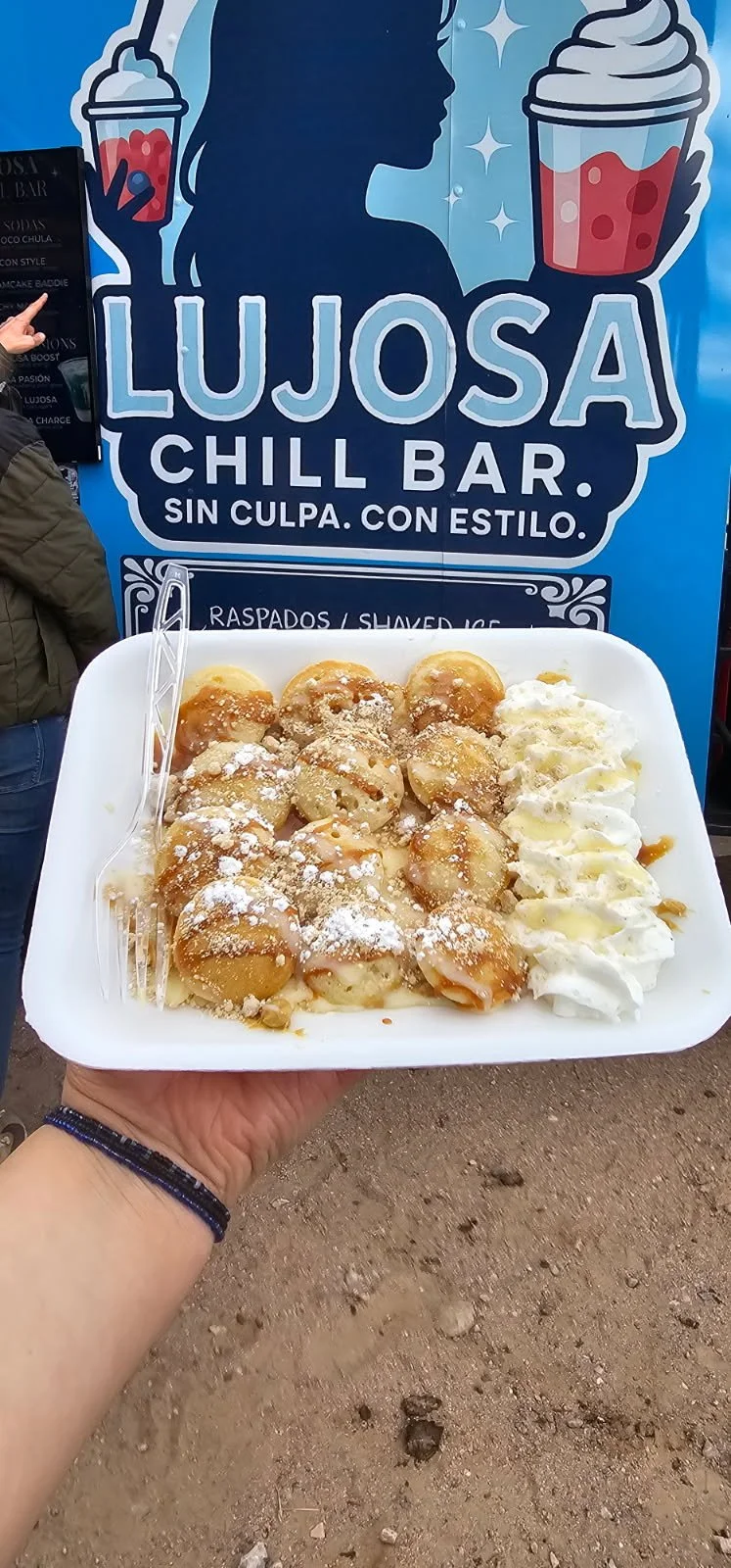 Hand holding a white tray of small cream-filled pastries topped with powdered sugar and drizzle, with whipped cream on the side, in front of a blue sign for Lujos Chill Bar.