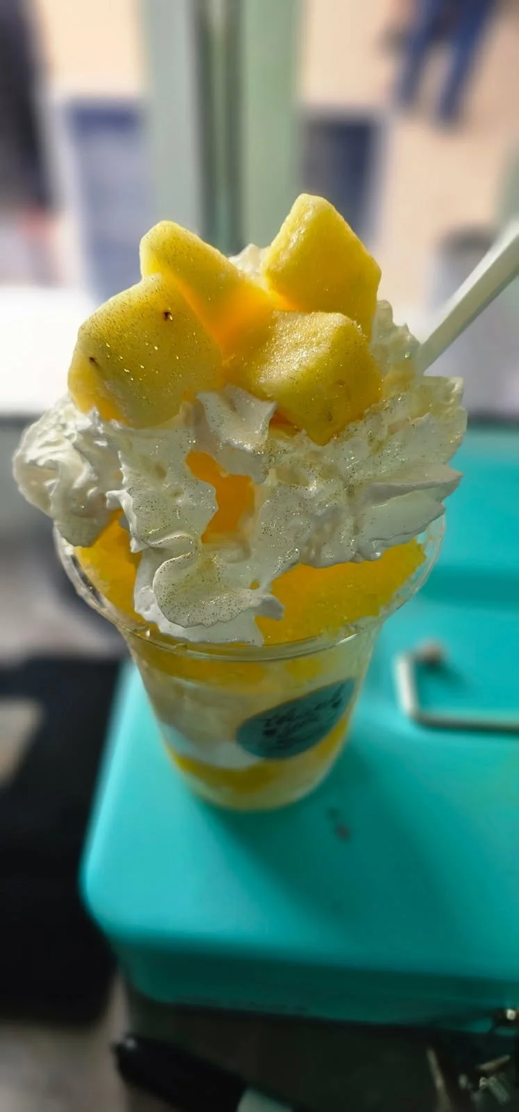 Mango-flavored raspado t with whipped cream and mango chunks in a clear plastic cup.