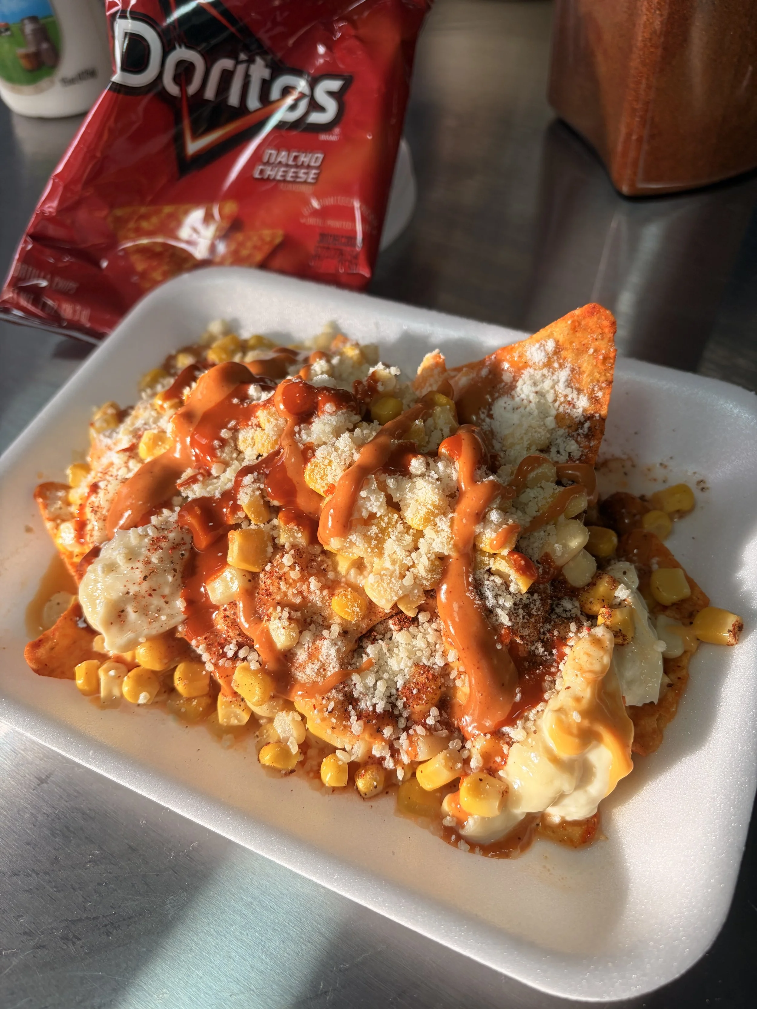 A plate of nachos topped with cheese, corn, and hot sauce, with a bag of Doritos Nacho Cheese flavored chips in the background.