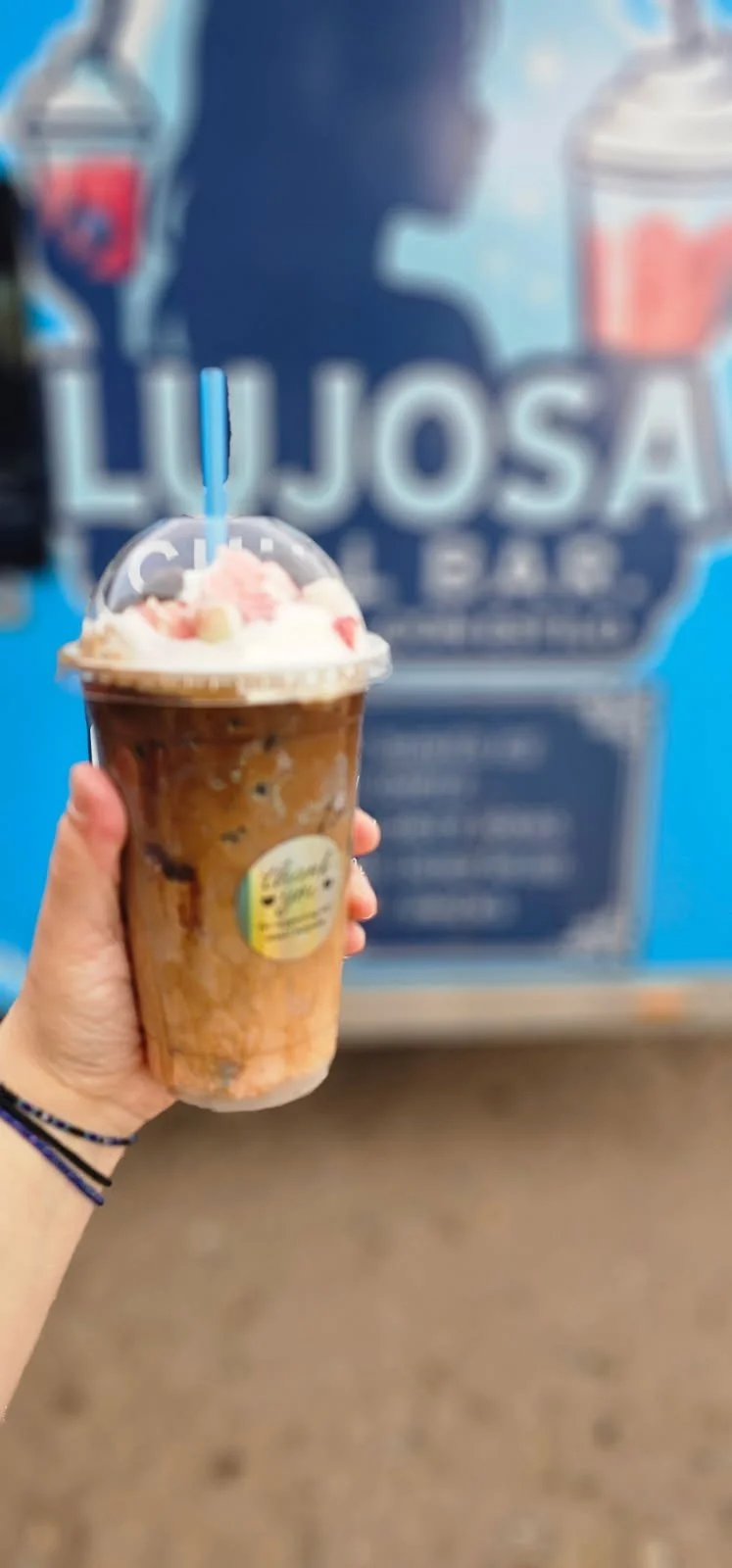 A hand holding a chocolate frosted milkshake with whipped cream and pink toppings in a clear plastic cup with a blue straw in front of a blue storefront with large sign reading 'LUJOSA' and a silhouette of a person with a beverage.