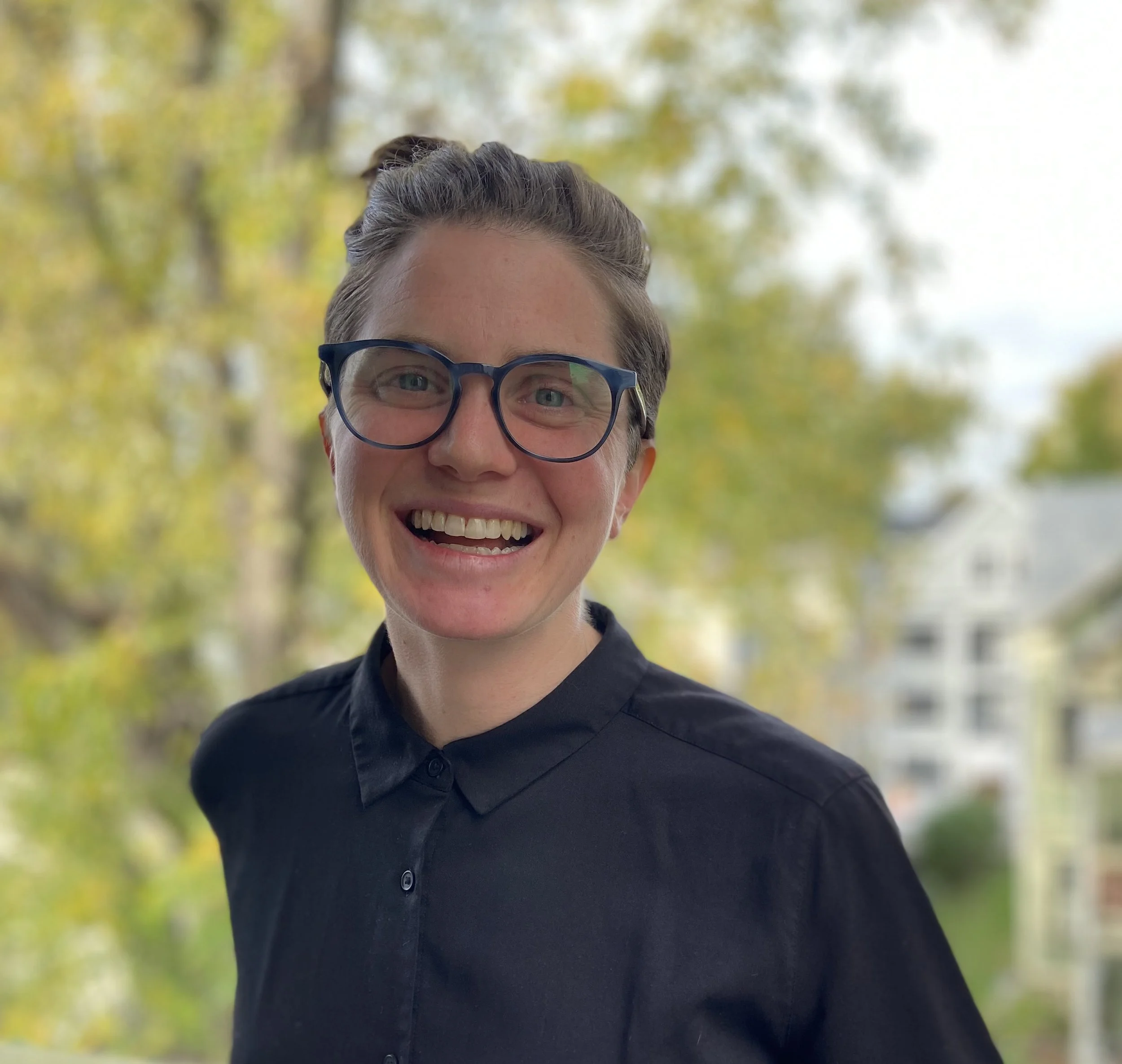 Michele wears a black button up and black framed glasses and stands in front of a tree in the background