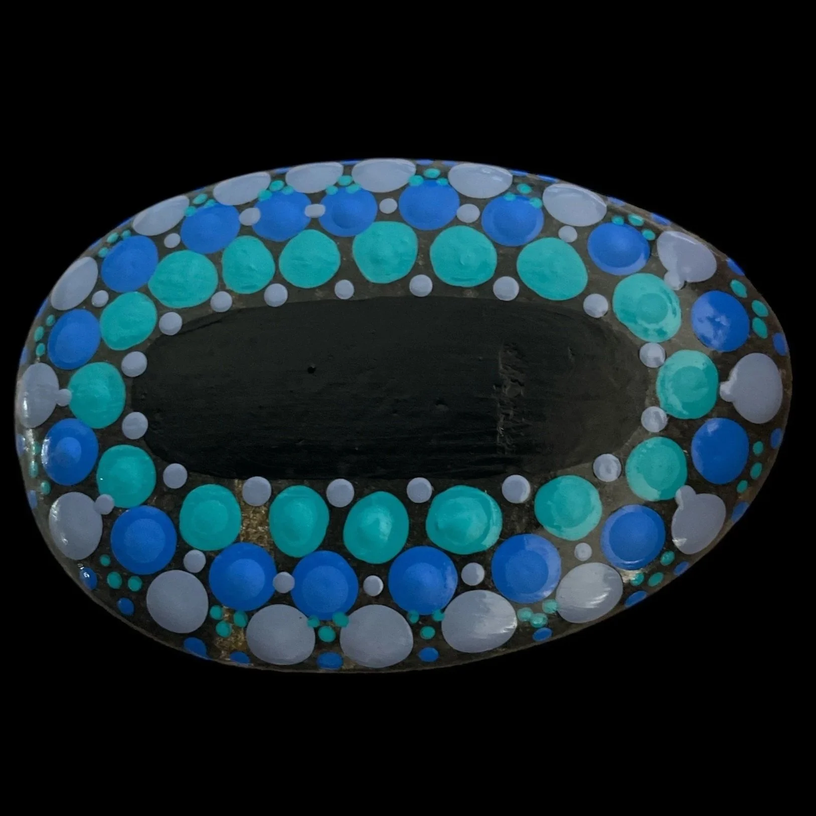 Oval rock painted with concentric dot pattern in blue, turquoise, and light blue on black background, set against solid black backdrop.