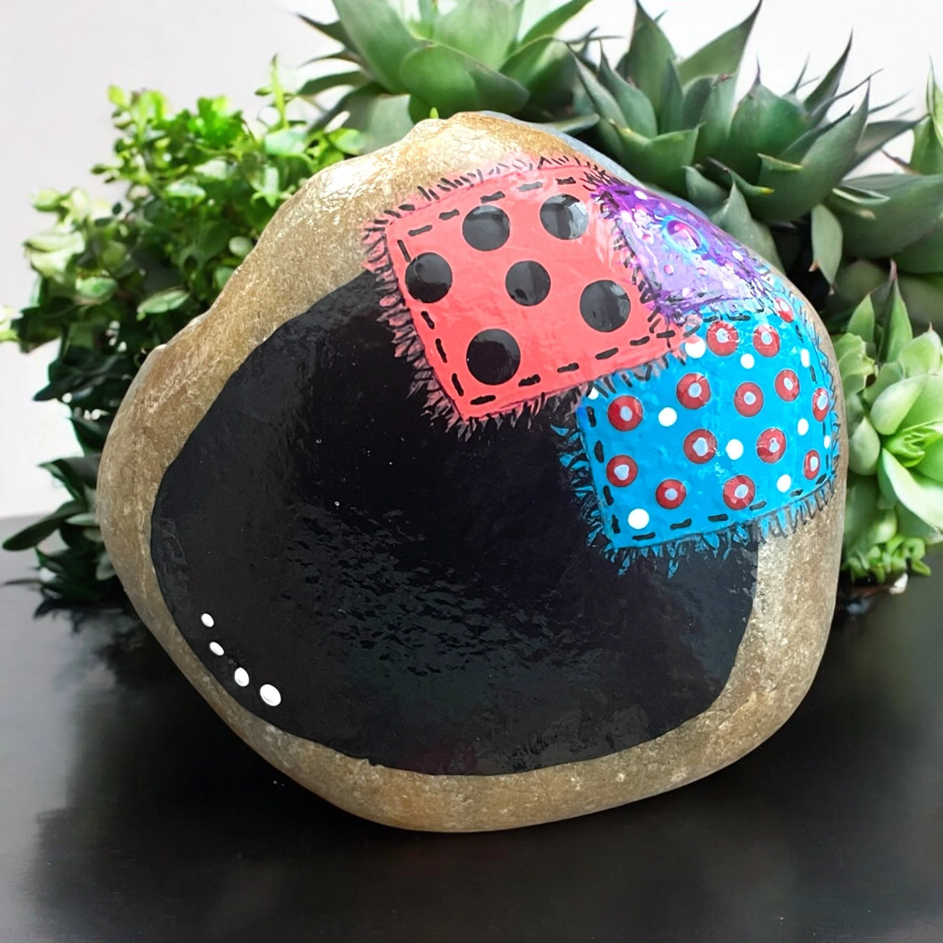 Painted rock of a black chalkboard with a colorful, patterned quilt patches, surrounded by succulents.