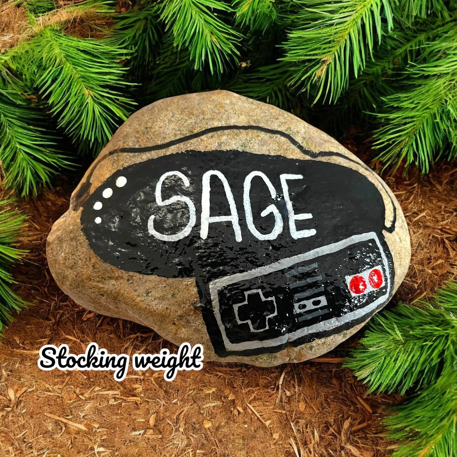A painted rock featuring the word 'SAGE' inside a black oval, a retro video game controller, and the words 'Stocking weight' at the bottom, surrounded by green pine branches and brown soil.