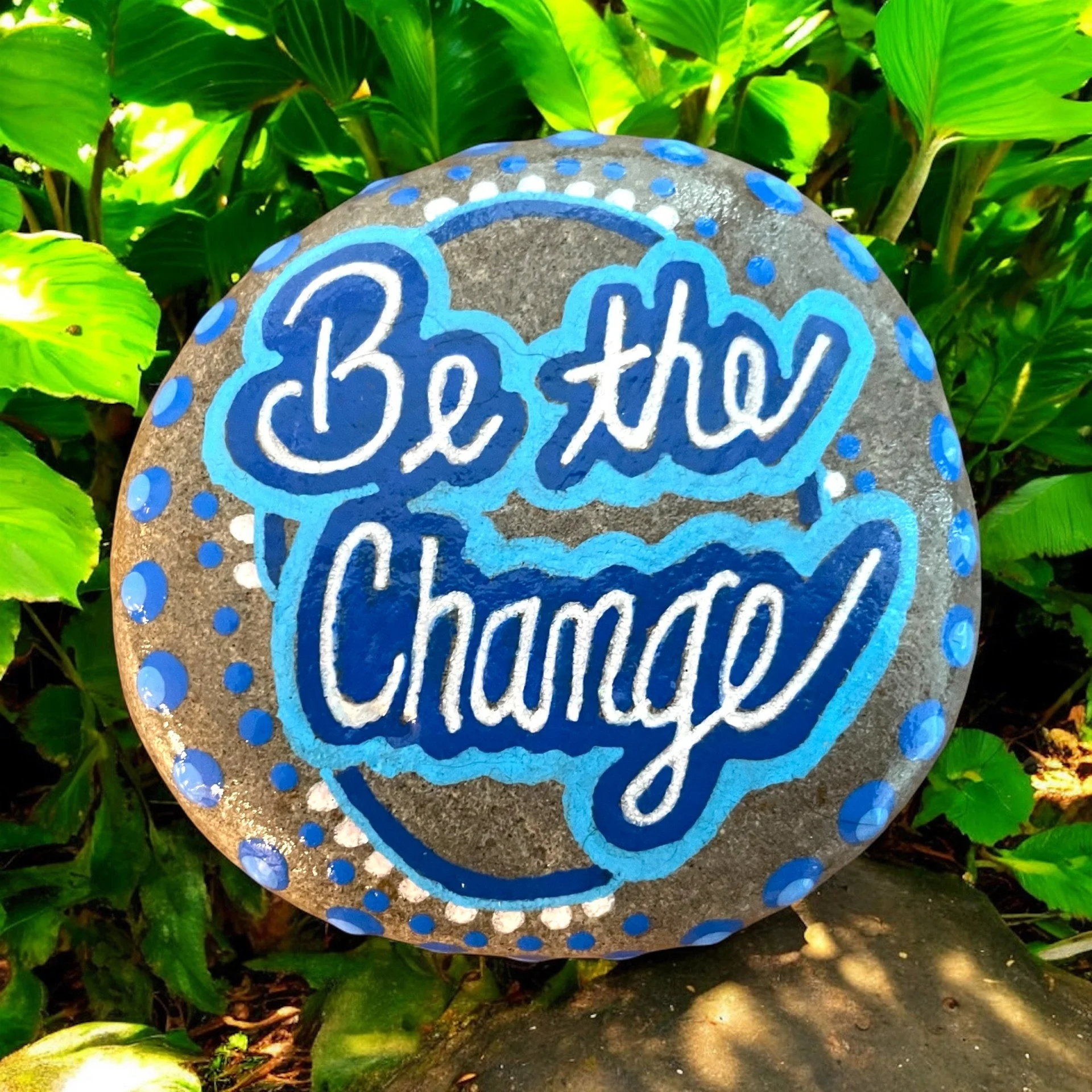 Oval rock painted with 'Be the Change' in bold cursive, outlined in blue and white on gray background with colorful blue dots.